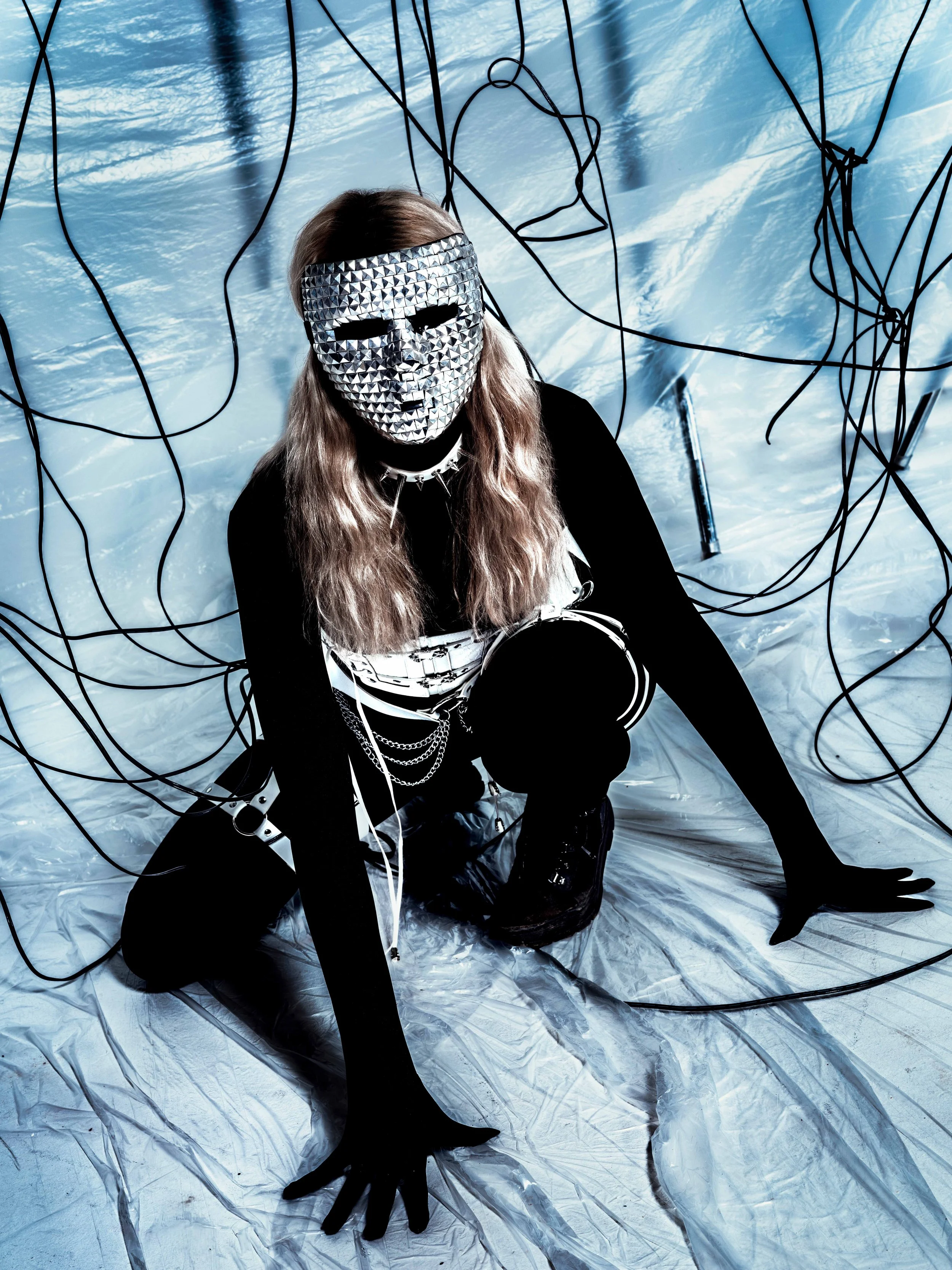 Person wearing a silver, studded mask and black clothing, crouching on reflective silver ground with black wires hanging overhead.