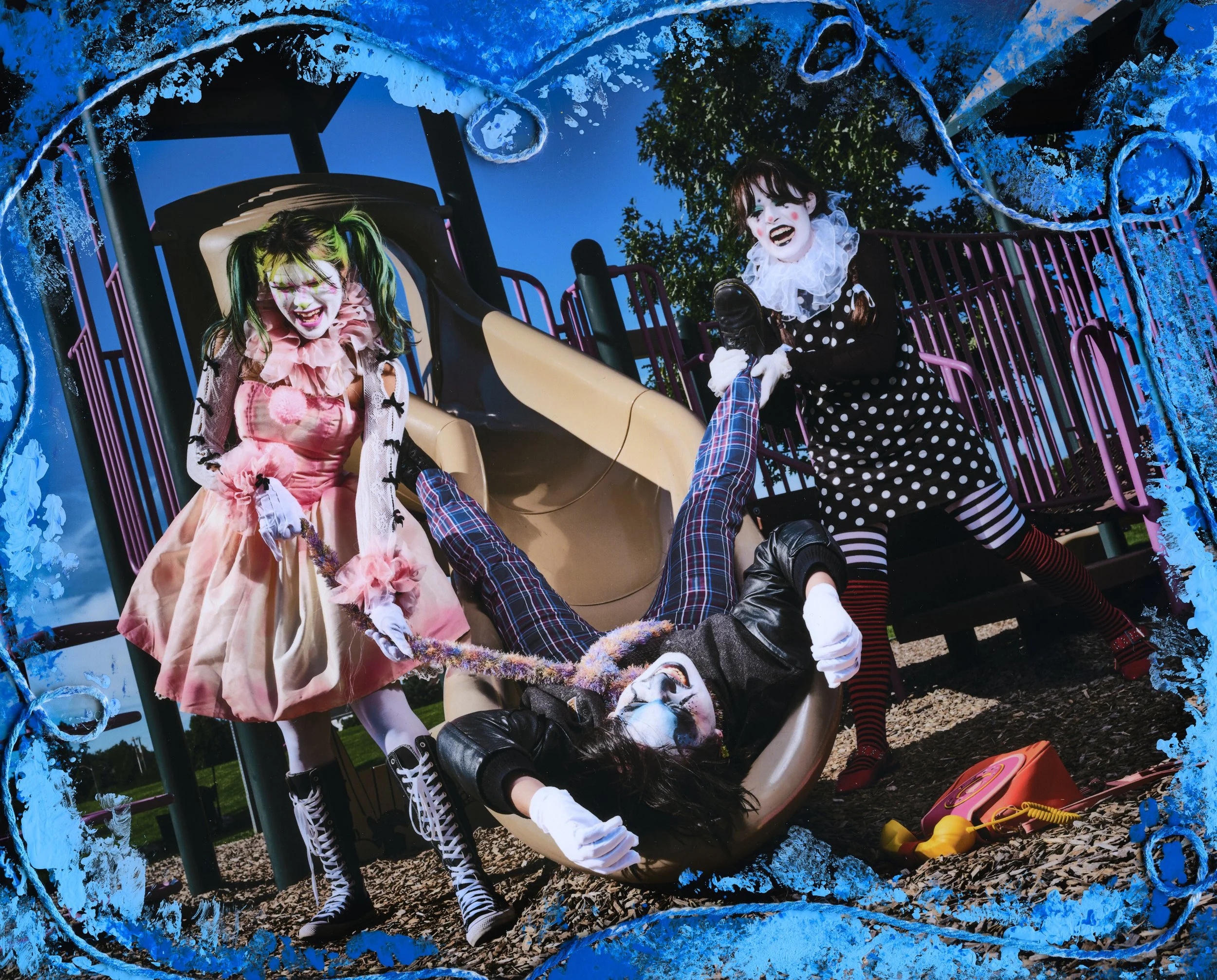 Two women dressed as clowns play on a playground slide during daytime. They are wearing exaggerated makeup, colorful costumes, and striped stockings. The scene is outdoors with a blue sky and green trees in the background. The image has a blue painted border.