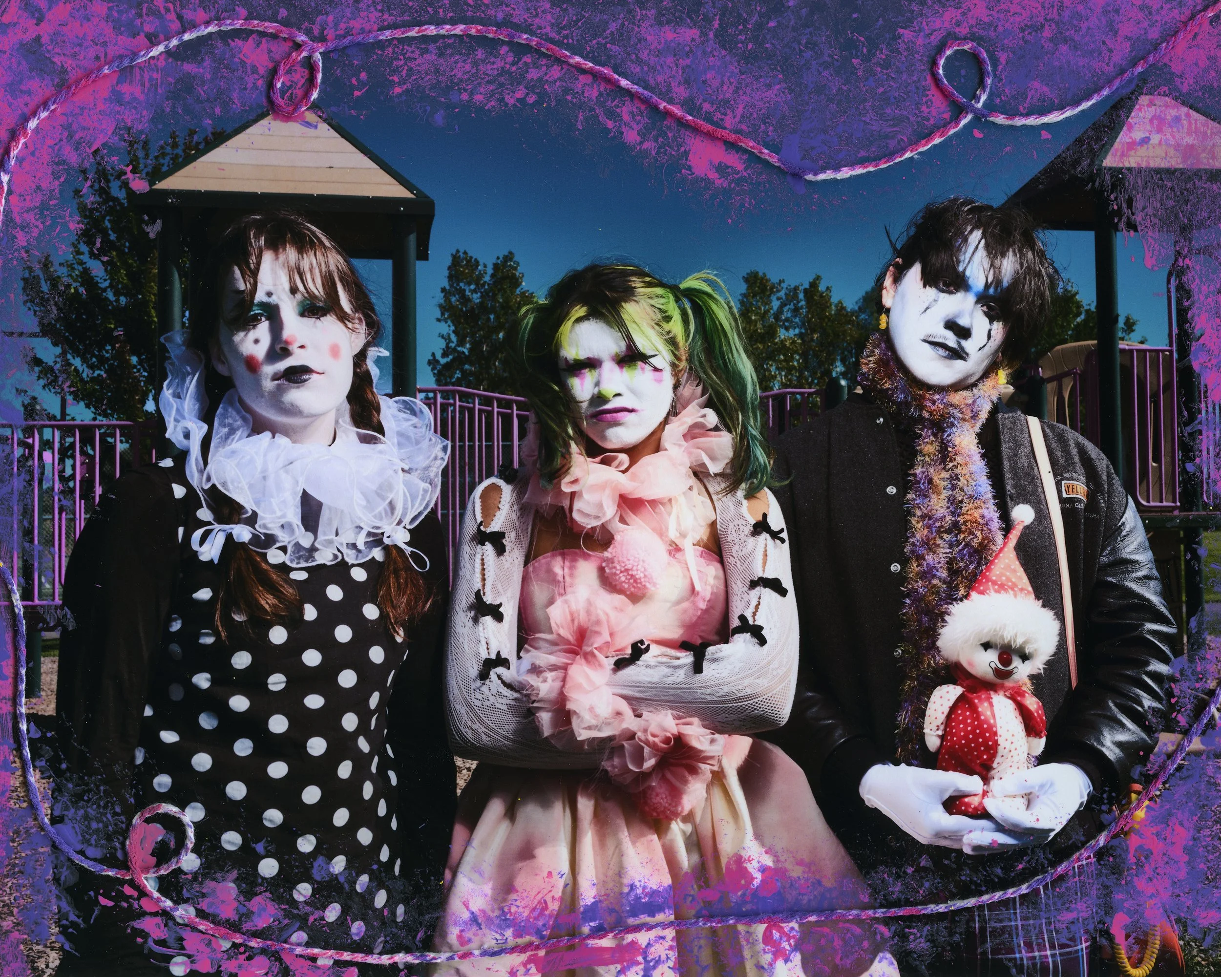 Three people dressed as clowns standing in front of a playground with trees in the background, with a decorative purple and pink border surrounding the image.
