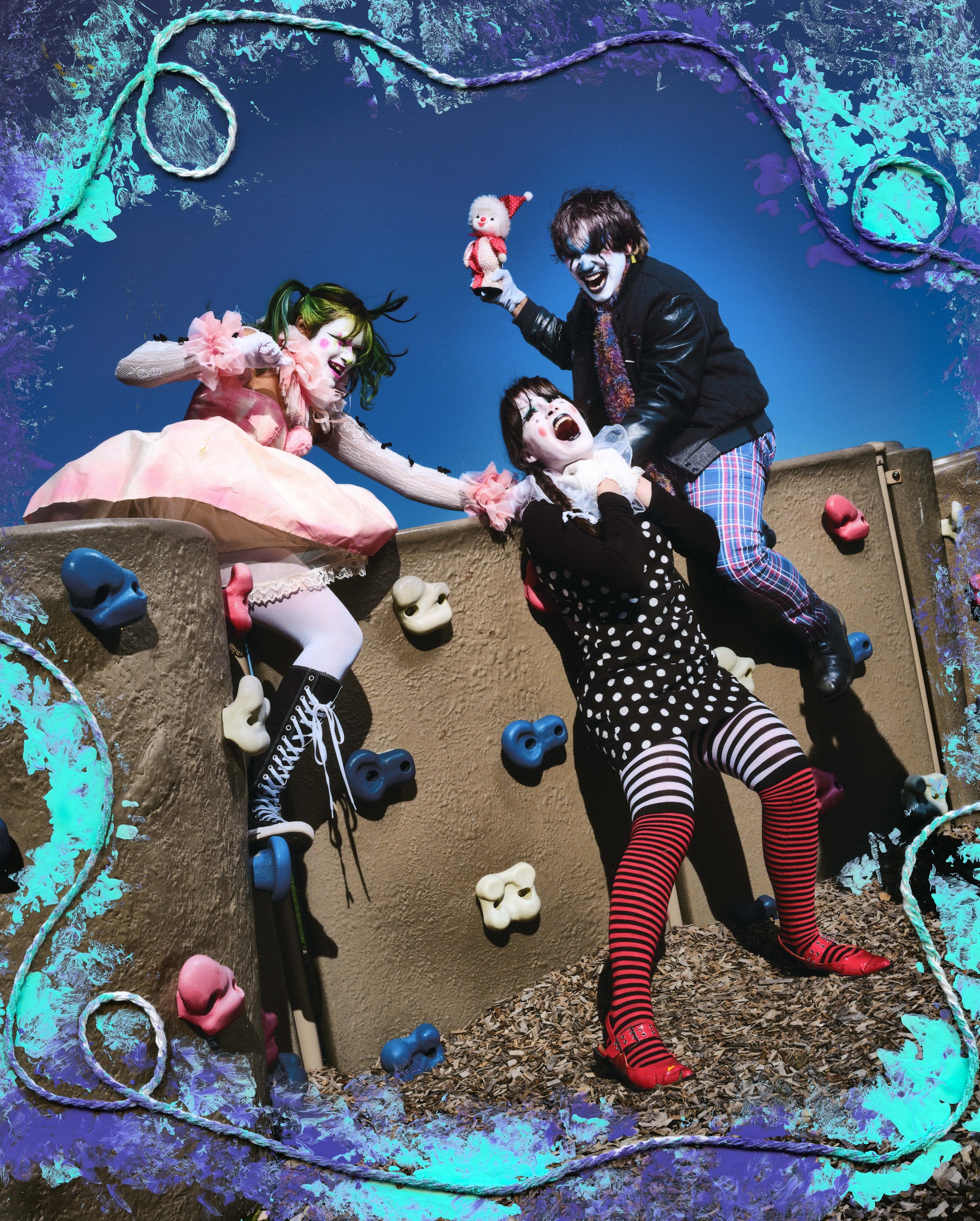 Three people dressed as clowns playing on a colorful climbing wall with skull-shaped hold decorations. One is holding a small doll with a red hat, another is dressed in a pink and white outfit with green hair, and the third is wearing black and white polka dot clothing with striped tights.