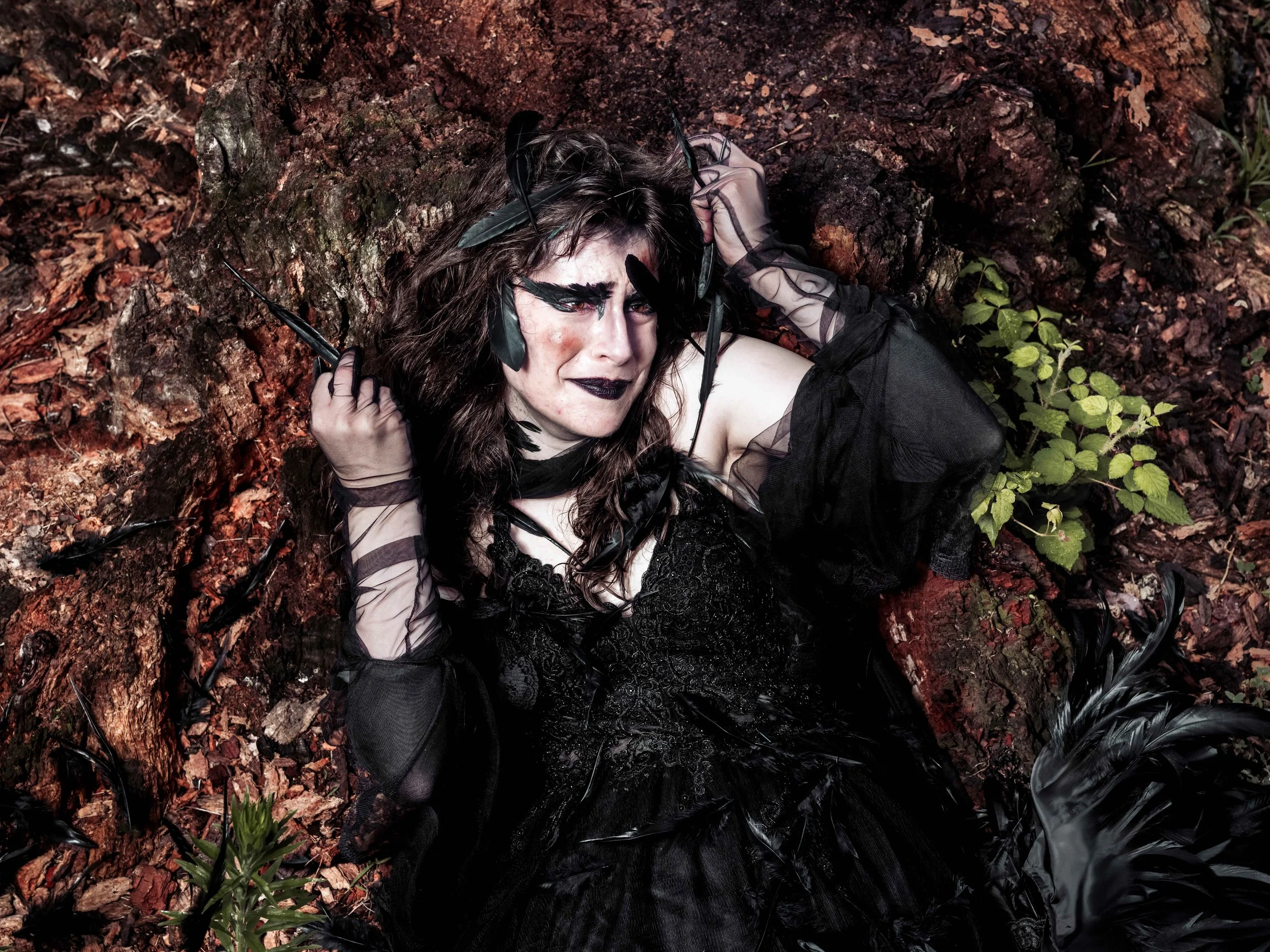 A woman with dark makeup and feather accessories lies on the ground in a forest, surrounded by leaves, soil, and small plants.