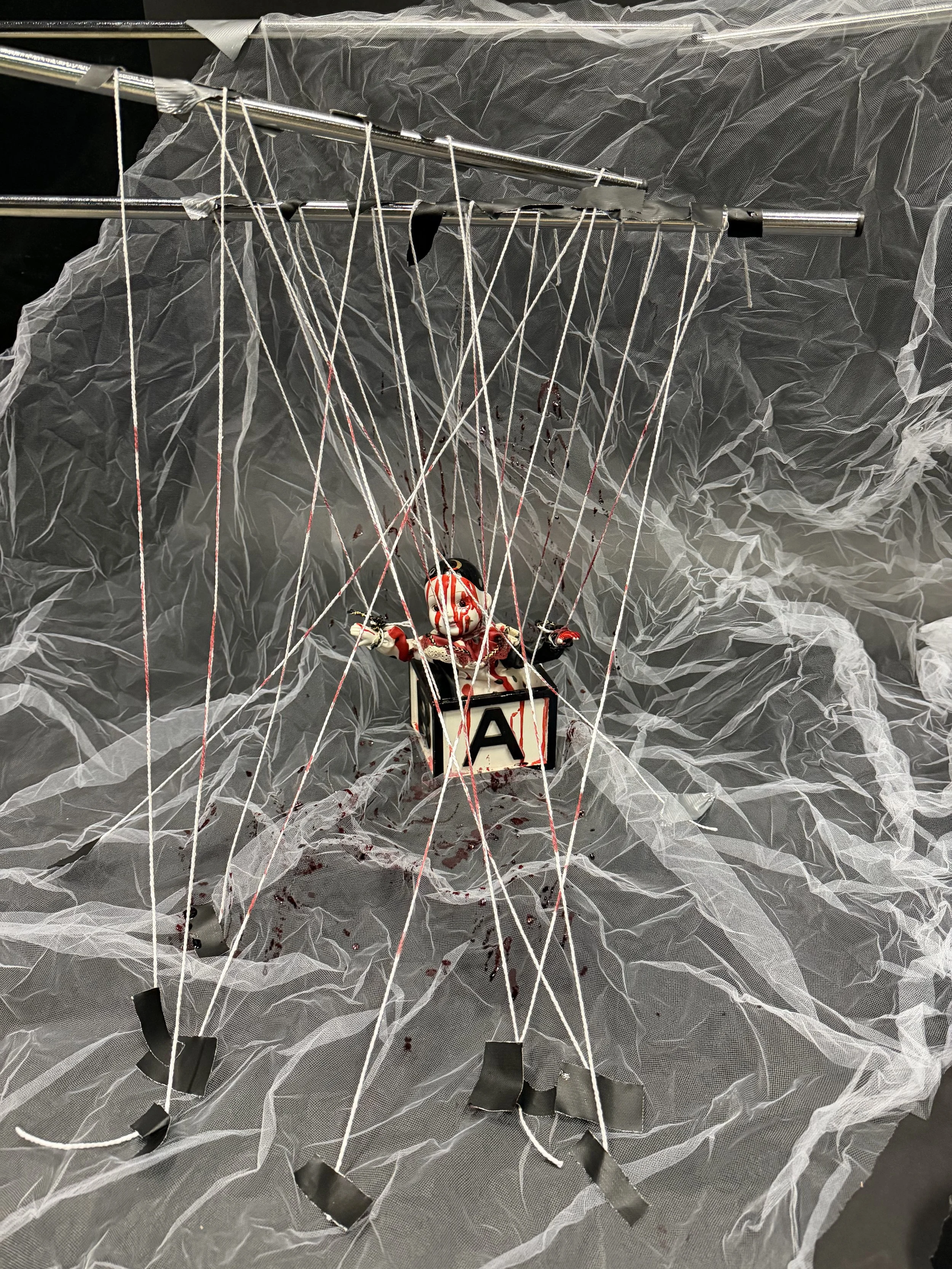 A doll with blood-streaked face and outfits hanging in black strings, set against a backdrop of crumpled, semi-transparent fabric that resembles a horror scene.