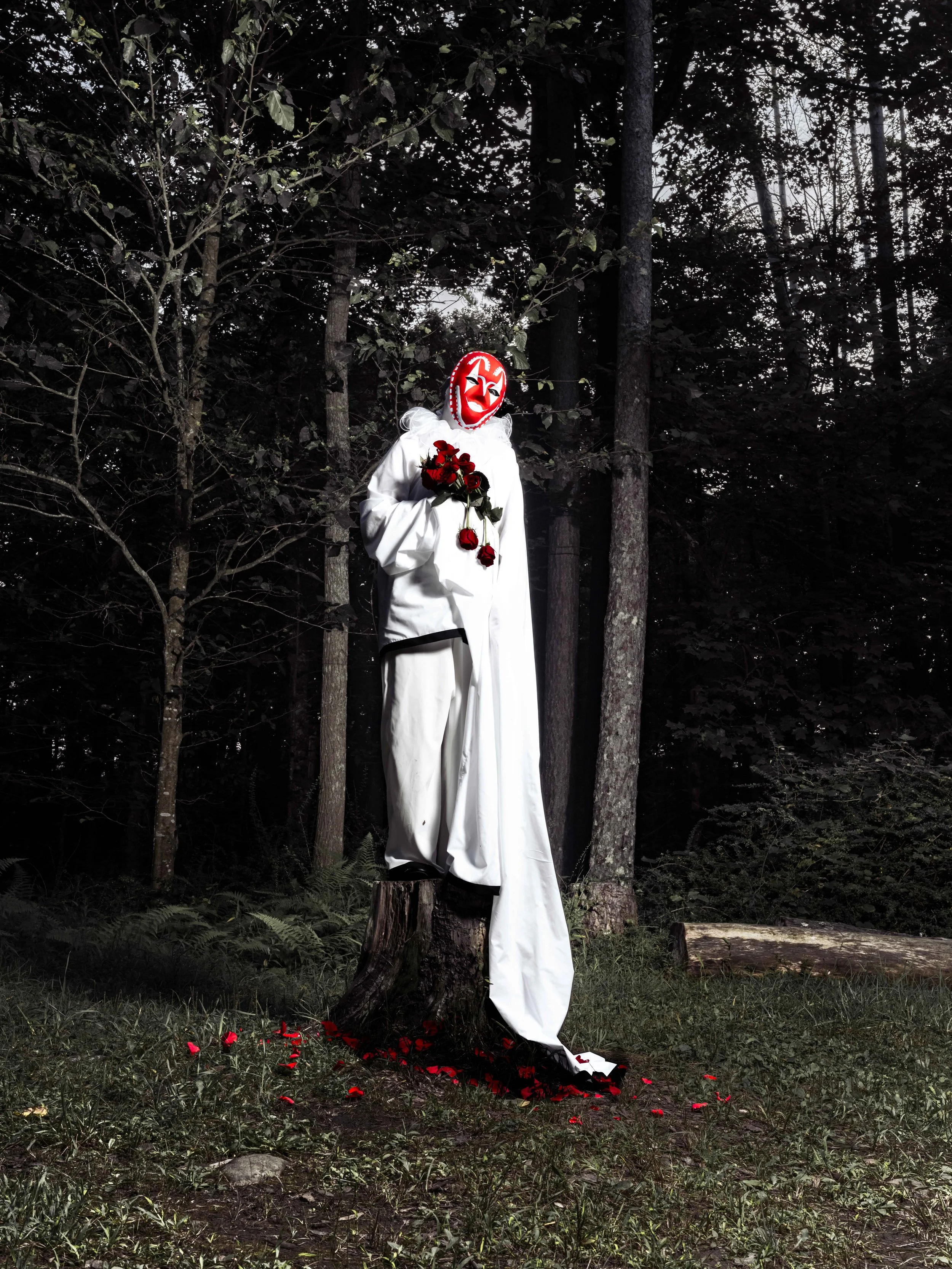 A scarecrow with a red clown mask, white clothing, holding red roses, standing in a wooded area at dusk with fallen petals on the ground.