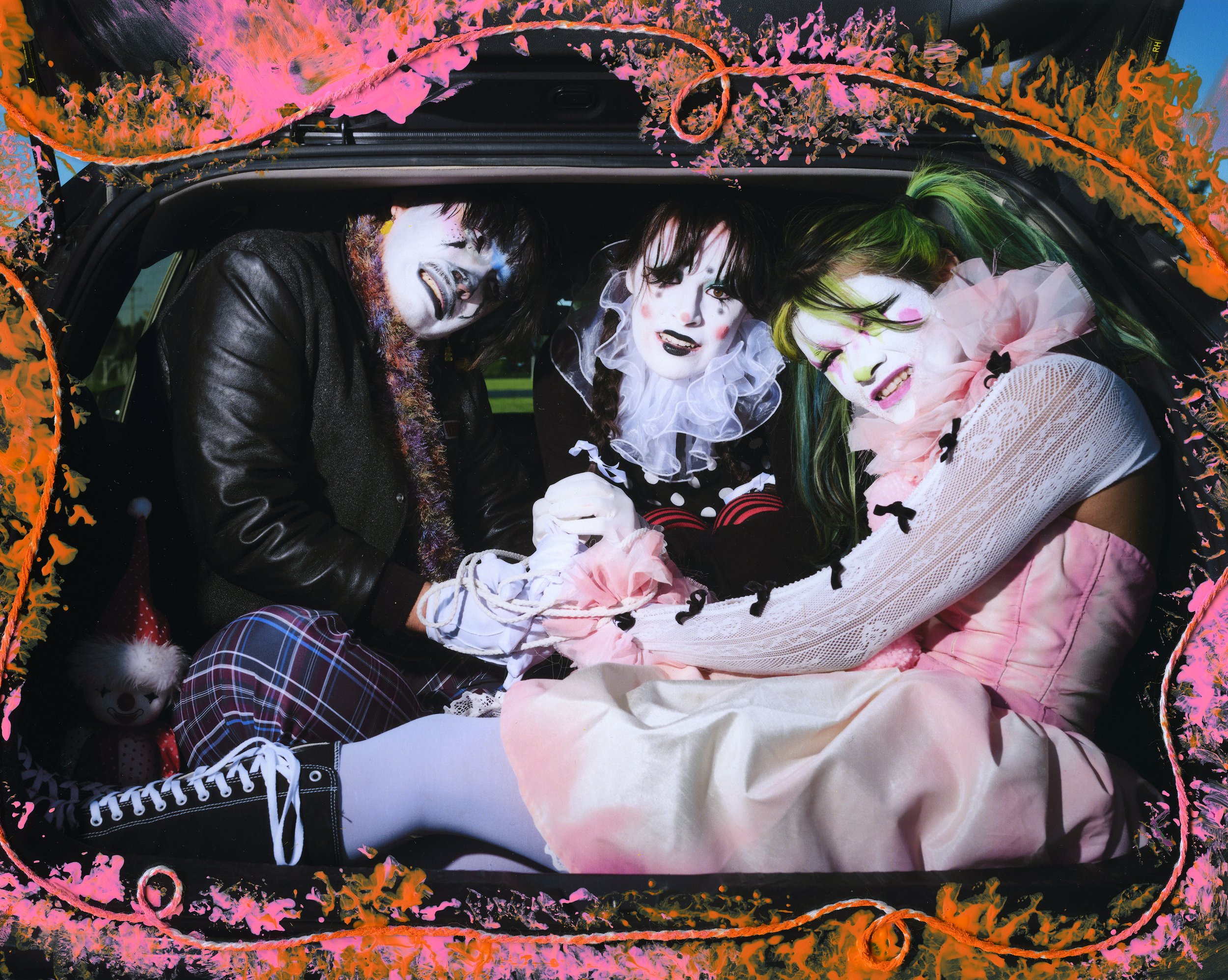Three individuals lying in the back of a decorated vehicle, dressed in clown or mime costumes with face paint, colorful hair, and costumes. The vehicle is decorated with pink and orange paint splashes and fabric.