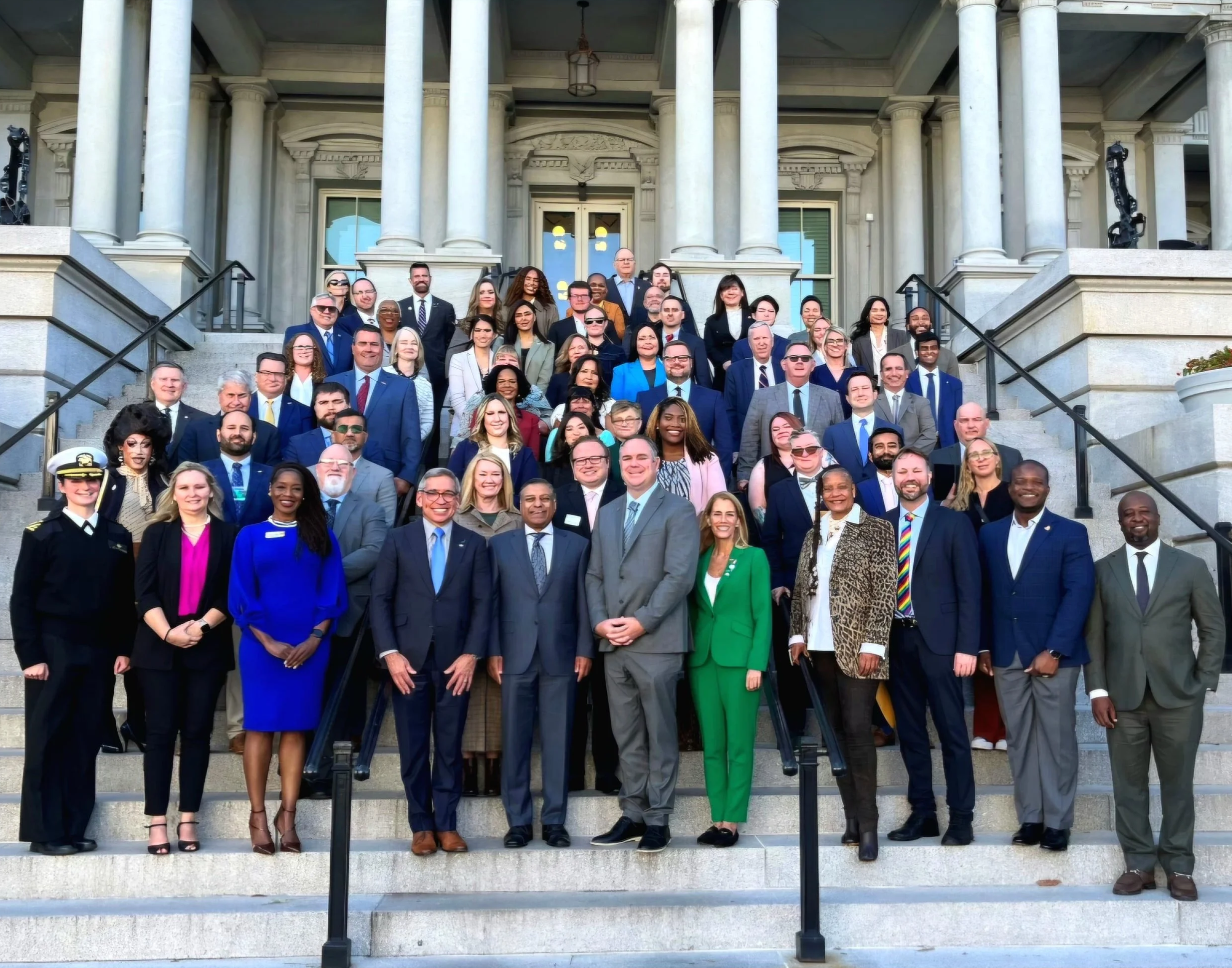 A large group of people gathered on the steps of a grand building with columns, posing for a group photo during daytime.