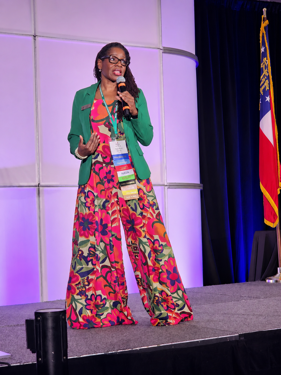 A woman speaking on stage at a conference, wearing a green blazer over a colorful floral dress, with conference badges hanging from her neck. There is an American flag to the right and purple lighting on the stage.