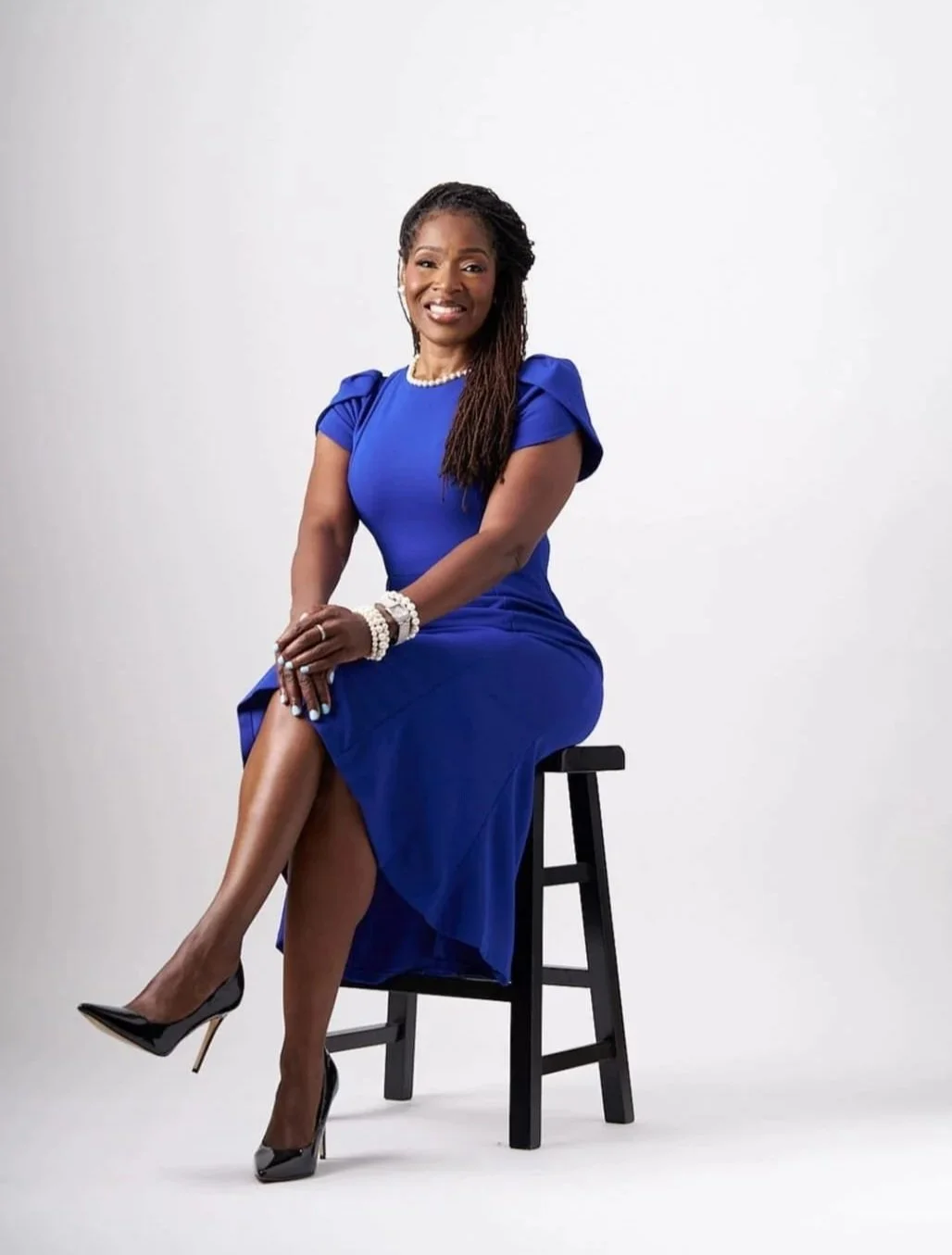 A woman in a royal blue dress sitting on a black stool with her legs crossed, wearing black high heels, pearl jewelry, and a smile, against a plain white background.