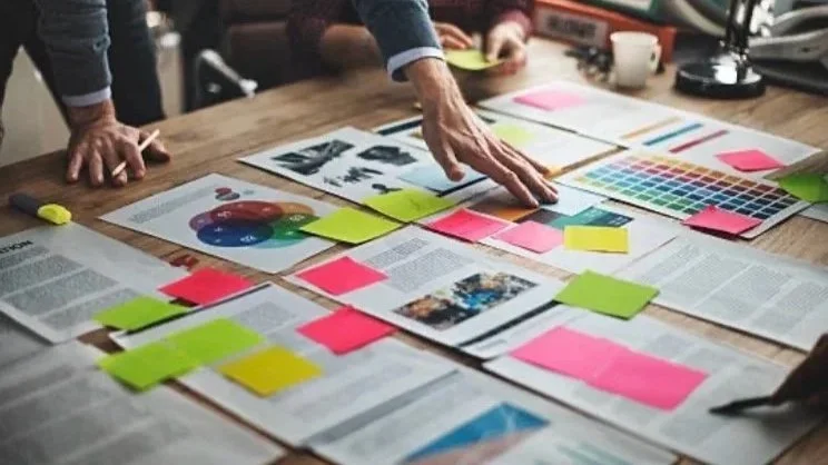 A person arranging documents, charts, and colorful sticky notes on a wooden table during a meeting or brainstorming session.