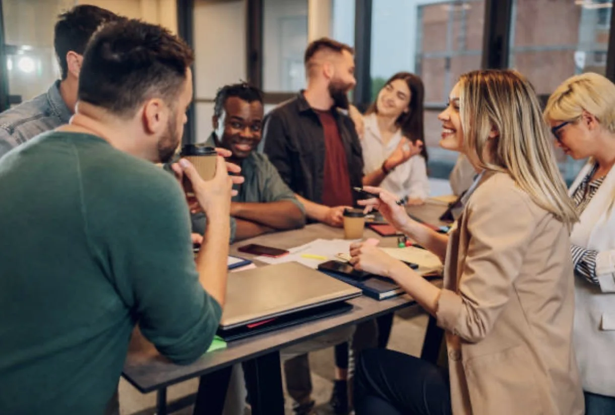 Group of diverse young adults gathered around a table in a modern office, smiling and engaging in conversation.