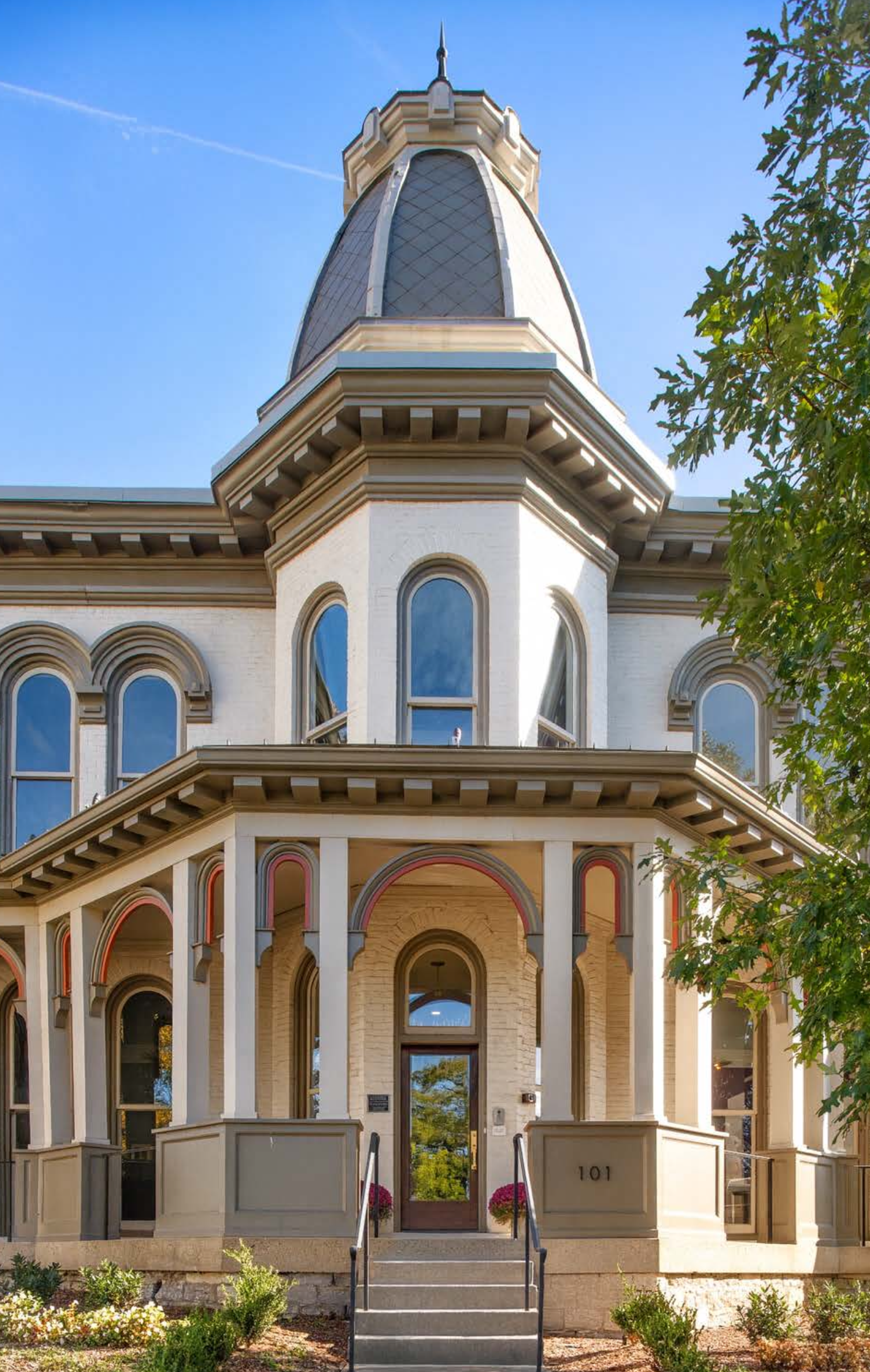 Front view of a historic Victorian house with a prominent tower, decorated with arched windows, and surrounded by a well-maintained garden.