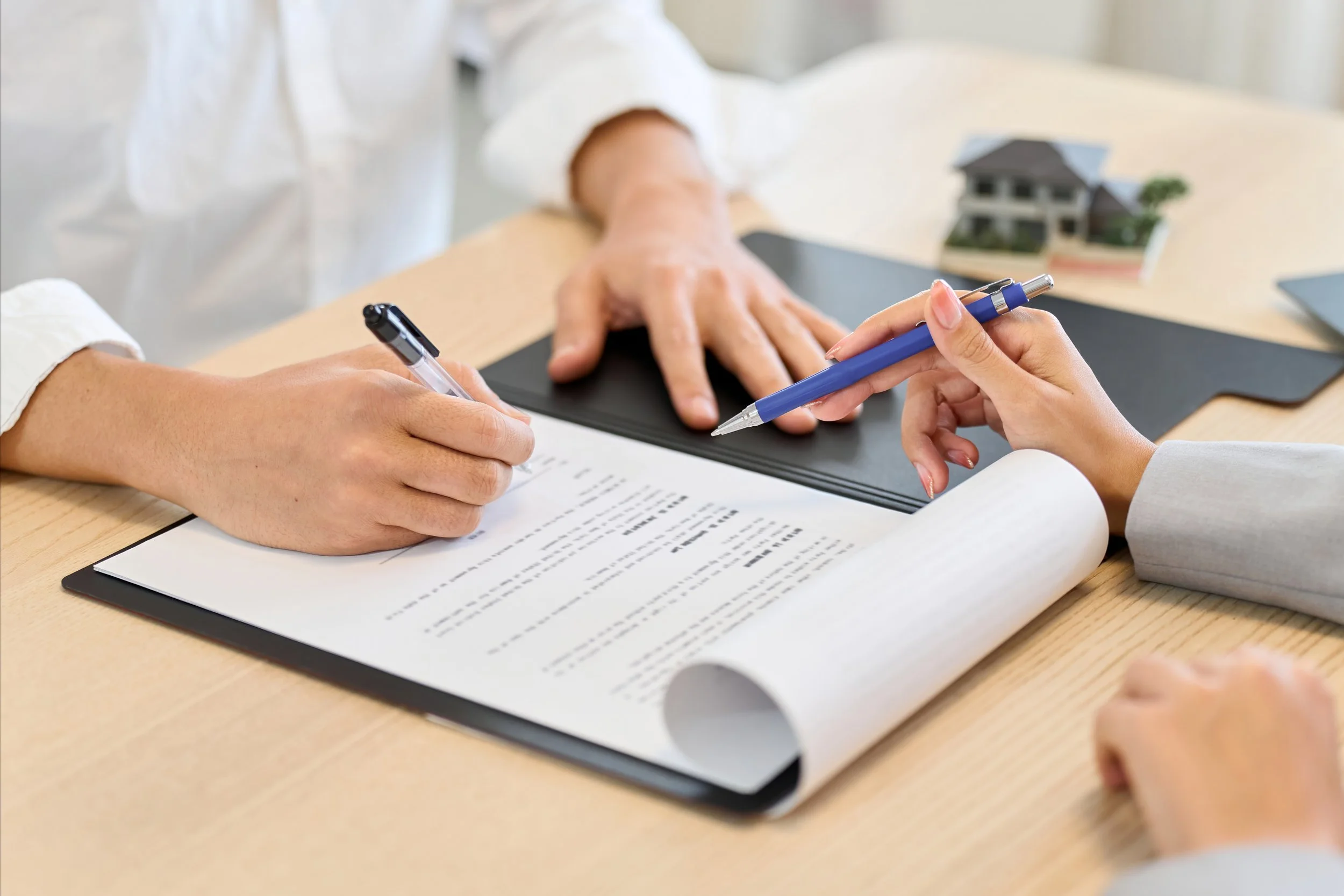 Two people sitting at a table reviewing and signing documents, one with a pen and the other using a pencil, with a small model house in the background.