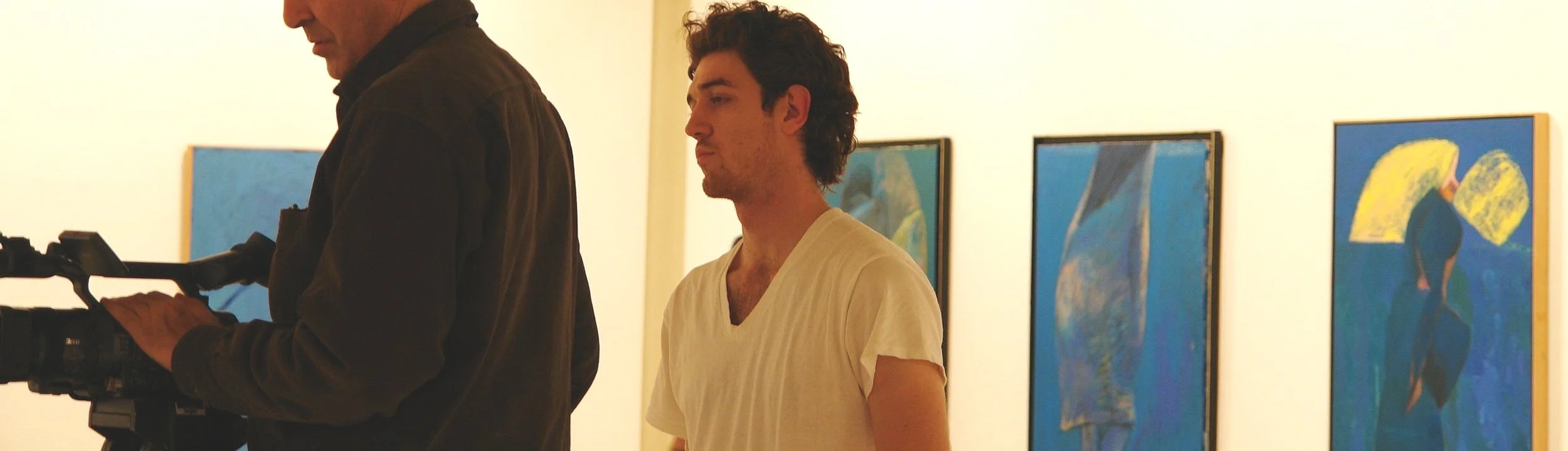Two men are standing in an art gallery, looking at paintings on the wall. One man is wearing a dark jacket and is operating a video camera, while the other man, with curly hair, is wearing a light-colored T-shirt.