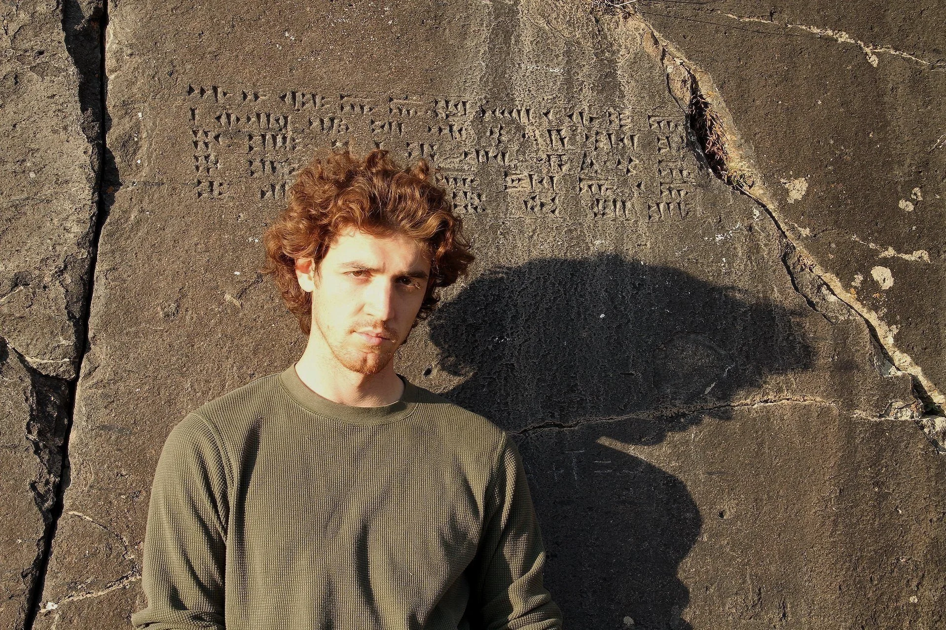 Young man with curly brown hair and a serious expression standing against a large weathered stone wall with ancient inscriptions. He is wearing an olive green long sleeve shirt, casting a shadow on the wall.