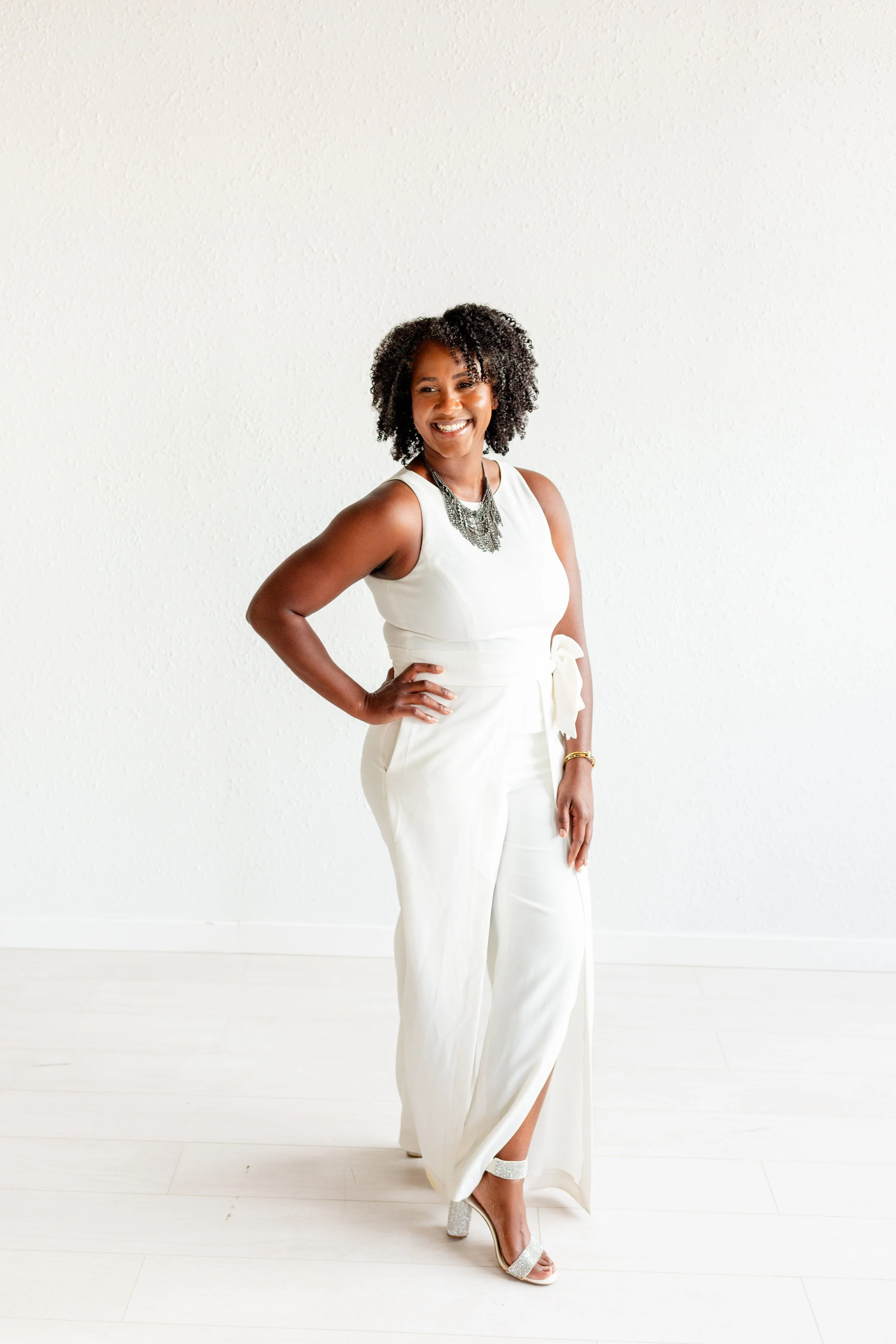 A woman with curly black hair smiling and standing against a plain white wall, wearing a white sleeveless dress with a bow at the waist, silver high-heeled shoes, and a black layered necklace.