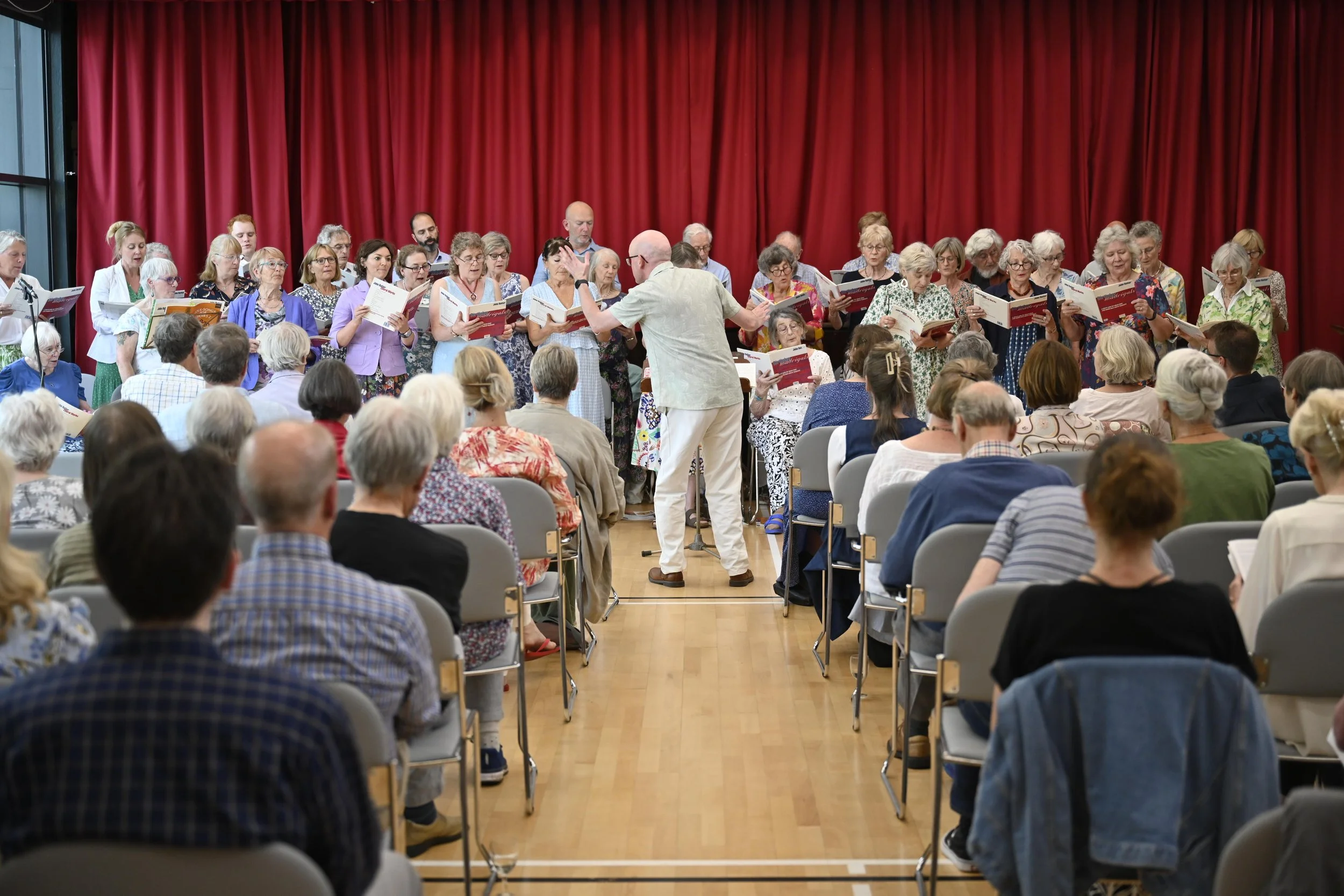 A choir of adults singing on stage during a performance in front of an audience, with a conductor leading them, set against red stage curtains.