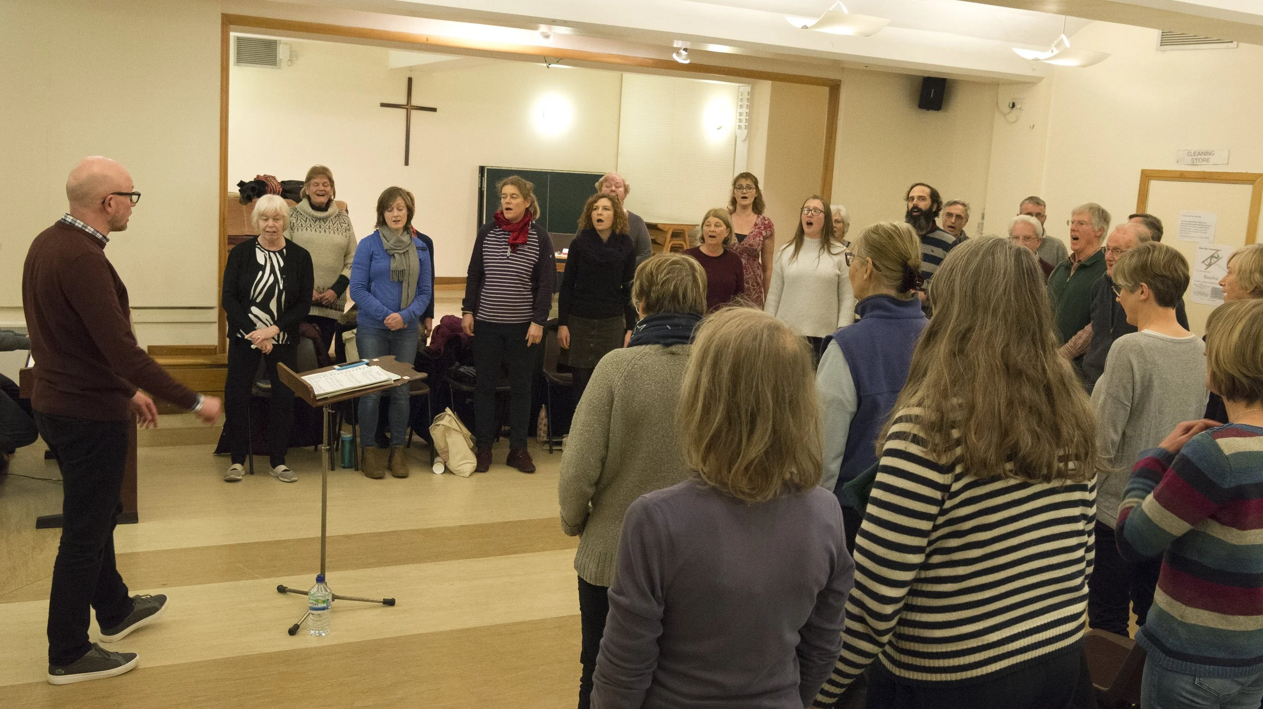 Group of people gathered indoors, singing or listening to a conductor in a church or community hall.