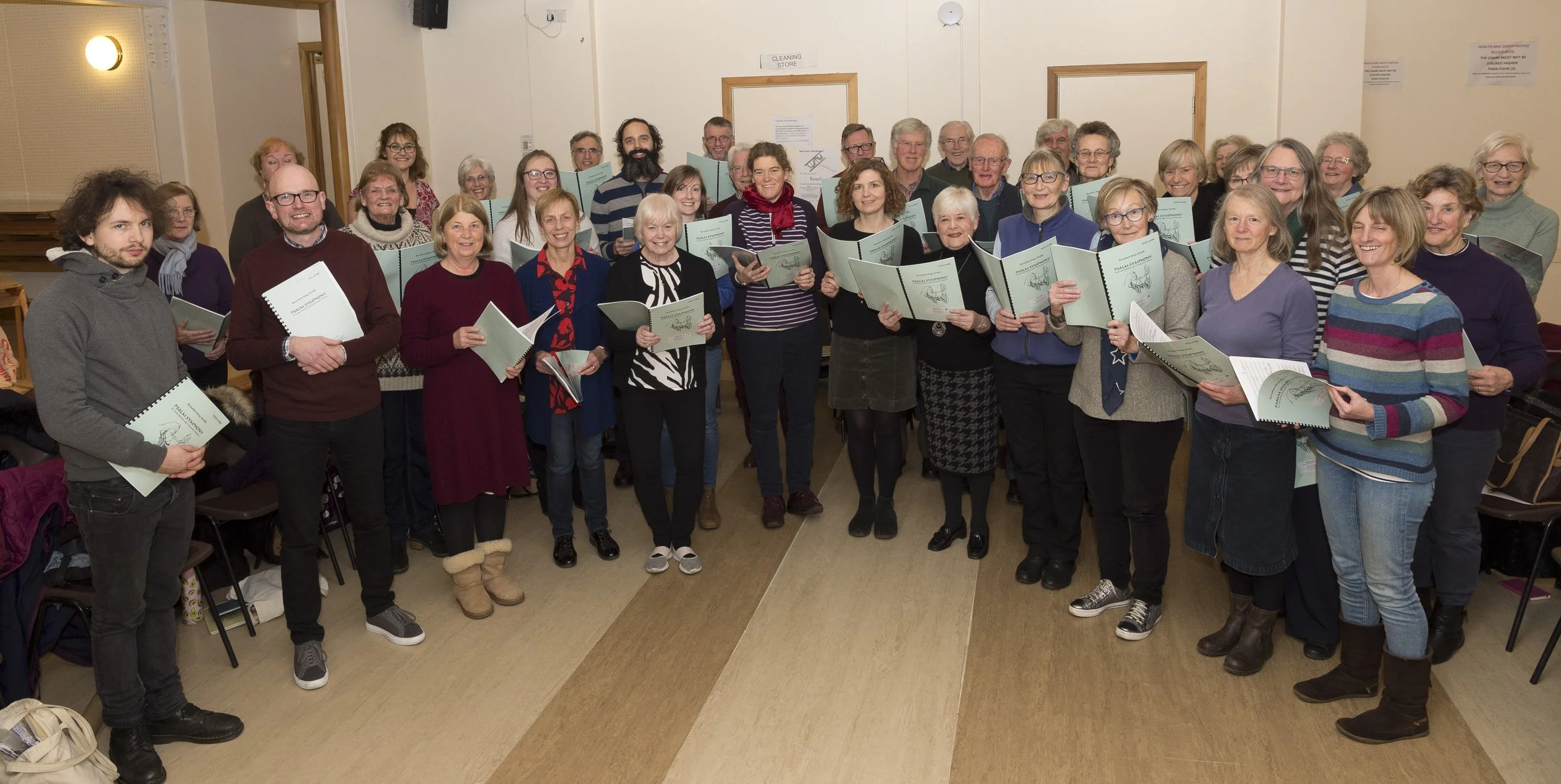 Group of people, about 35, standing in a room, holding music books, smiling at the camera.