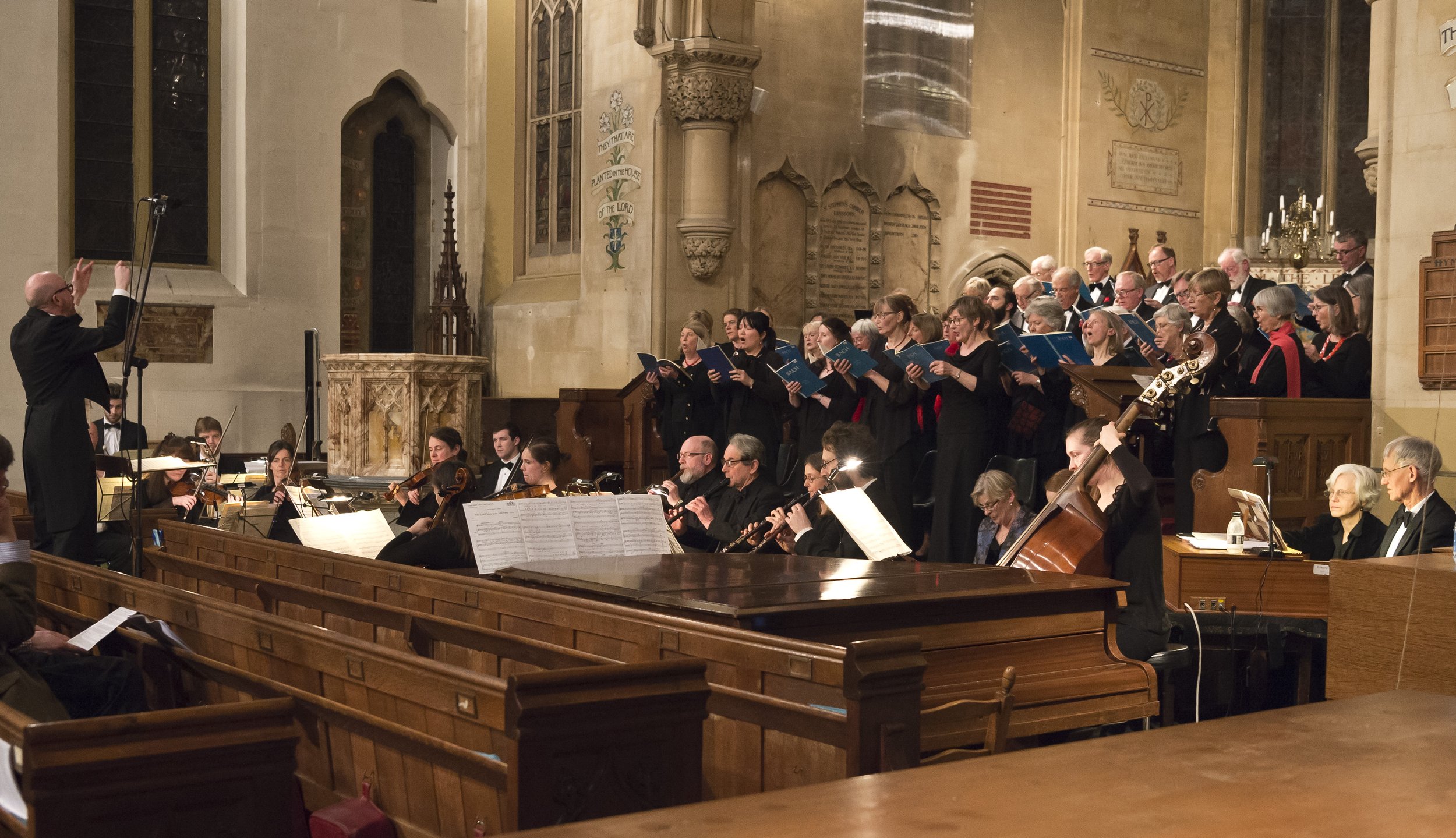 An orchestra and choir performing inside a church, with the conductor leading them, with stained glass windows and ornate decorations in the background.