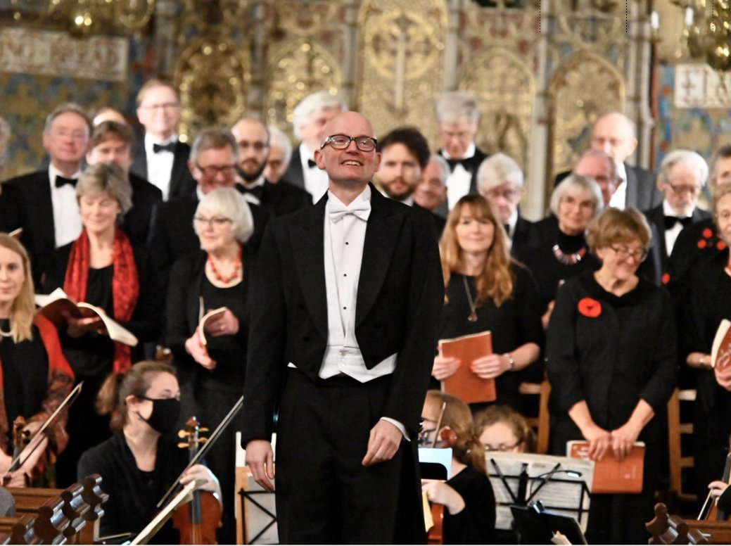 A male conductor in a tuxedo leading a choir and orchestra in a concert hall with ornate gold decorations.