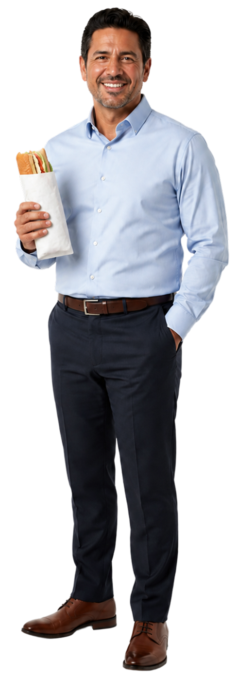 A man with dark hair and a beard smiling, wearing a light blue button-up shirt and dark pants, holding a sandwich wrapped in white paper in his right hand, standing against a plain background.