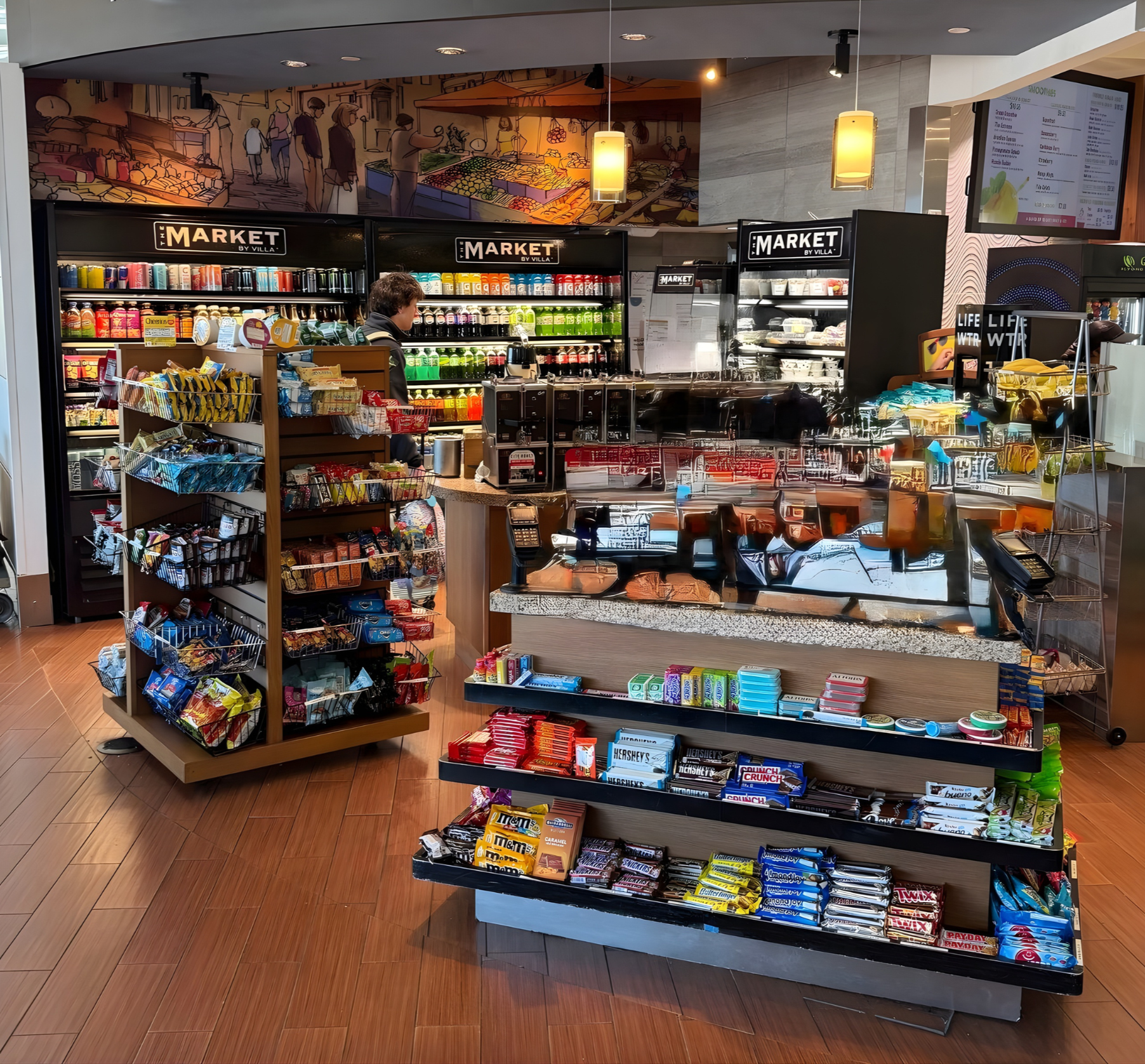 Inside a convenience store with shelves stocked with snacks, candies, and beverages. A customer is browsing near the checkout counter.