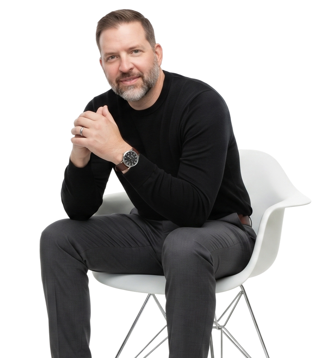 A man with a beard and mustache sitting on a white chair, wearing a black long-sleeve shirt, gray plaid pants, and a wristwatch, with his hands clasped together and smiling at the camera.
