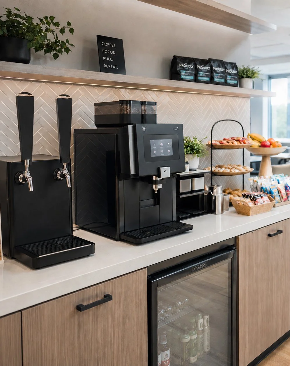 Coffee station with a coffee machine, two beverage taps, a variety of snacks, and a small refrigerator. Decor includes potted plants and a sign that reads 'Coffee. Focus. Fuel. Repeat.'