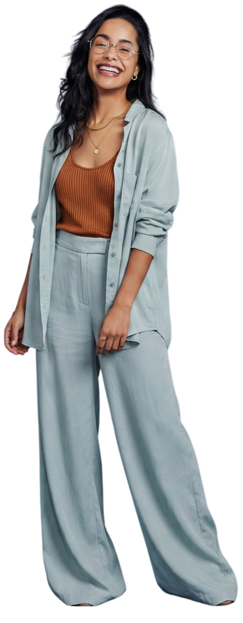 A woman with glasses, long black hair, and hoop earrings, smiling and standing with her hand at her side. She is wearing a light gray oversized shirt over a rust-colored sleeveless top and matching light gray wide-leg trousers. She accessorizes with a necklace with a pendant.