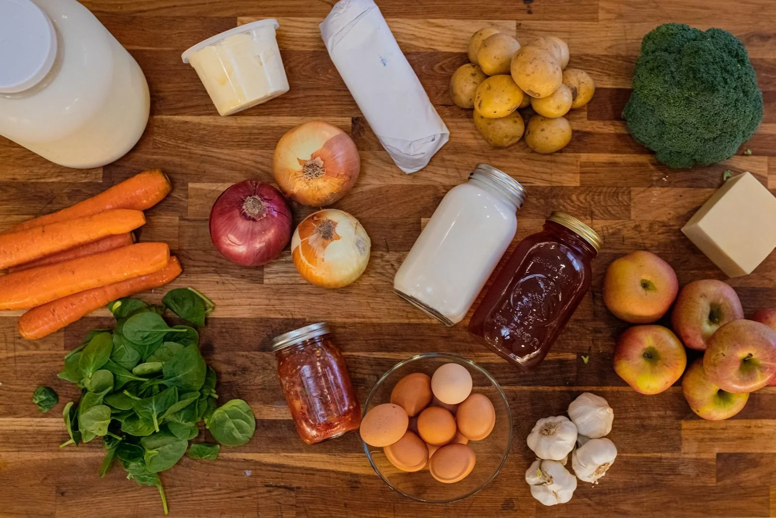 Assorted fresh vegetables, fruits, dairy, and eggs available through Moxie Farm's food membership. Items include carrots, onions, apples, potatoes, broccoli, garlic, eggs, cheese, butter, milk, and jars of jam and yogurt.