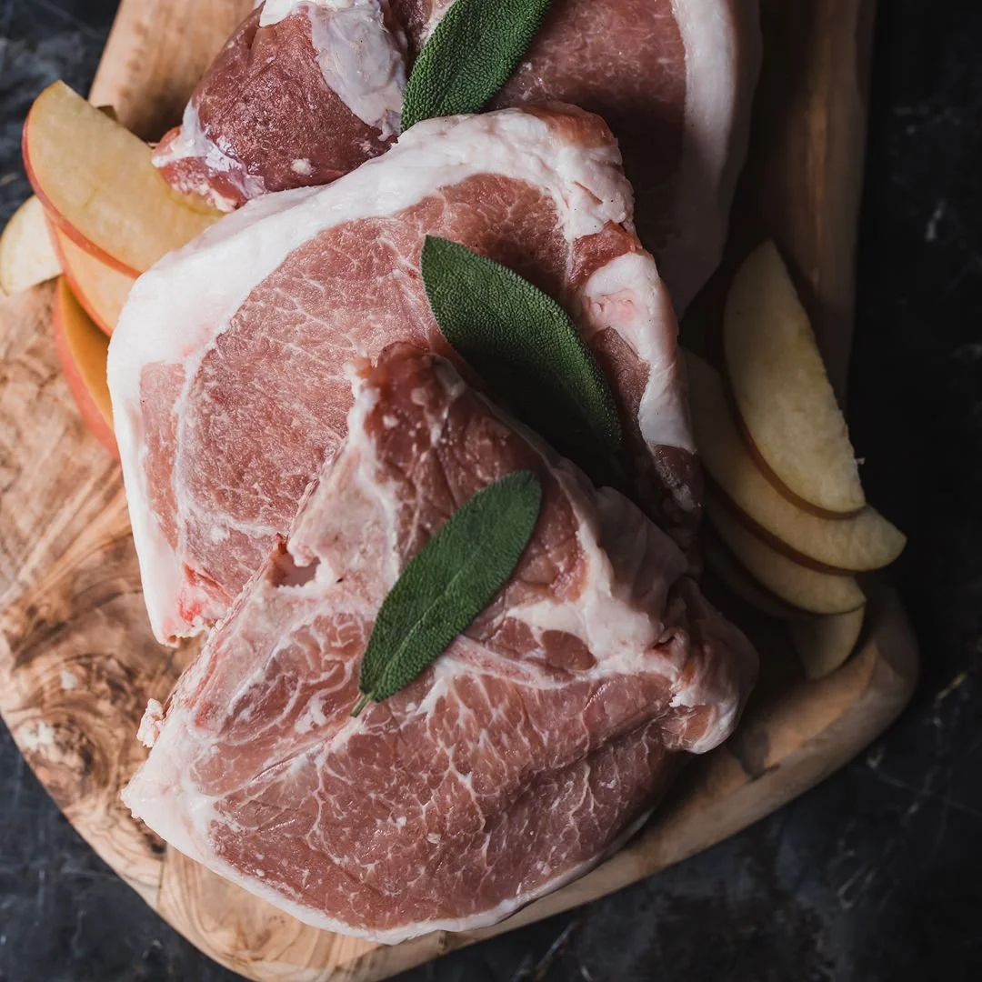 Raw pork chop with sage leaves, apple slices, and lemon wedges on a wooden cutting board.