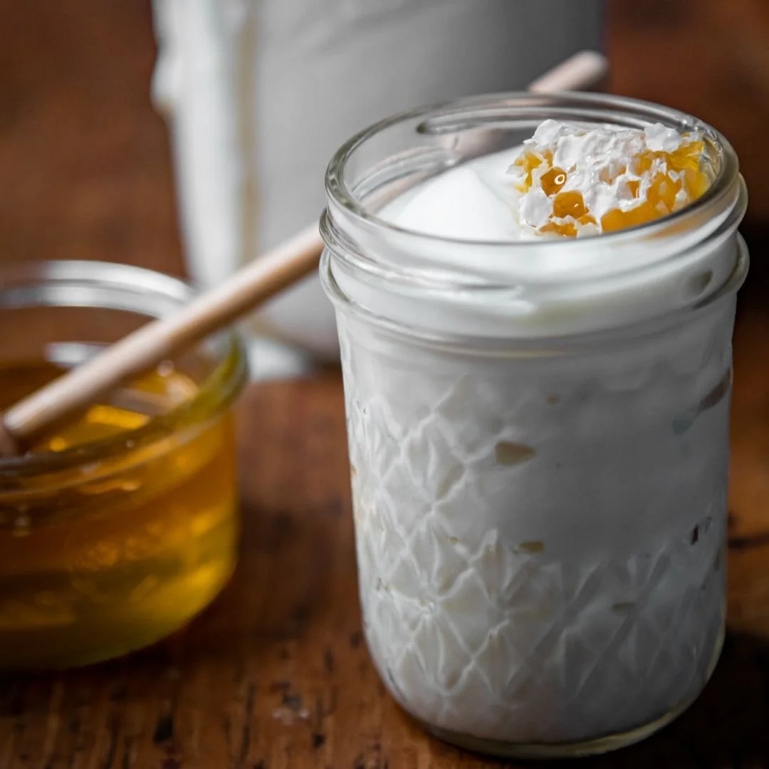 A glass jar filled with whipped cream topped with caramel sauce, next to a small bowl of honey and a wooden honey dipper on a wooden table.