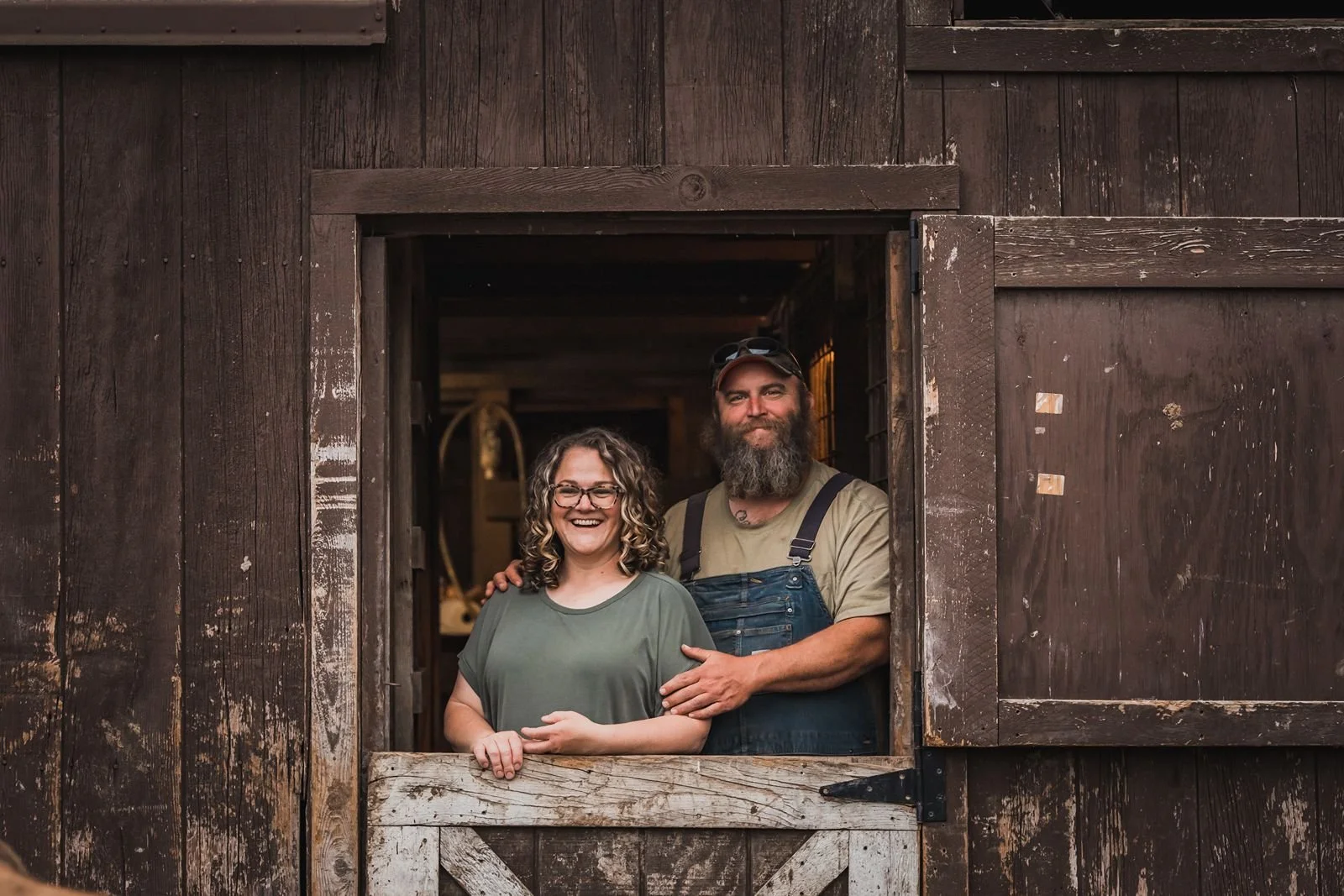 Matt and Jenny Vahdick standing in the door of the wooden barn at Moxie Farms. Jennifer is smiling and has curly hair and glasses; Matt is a a bearded man wearing sunglasses on his cap.