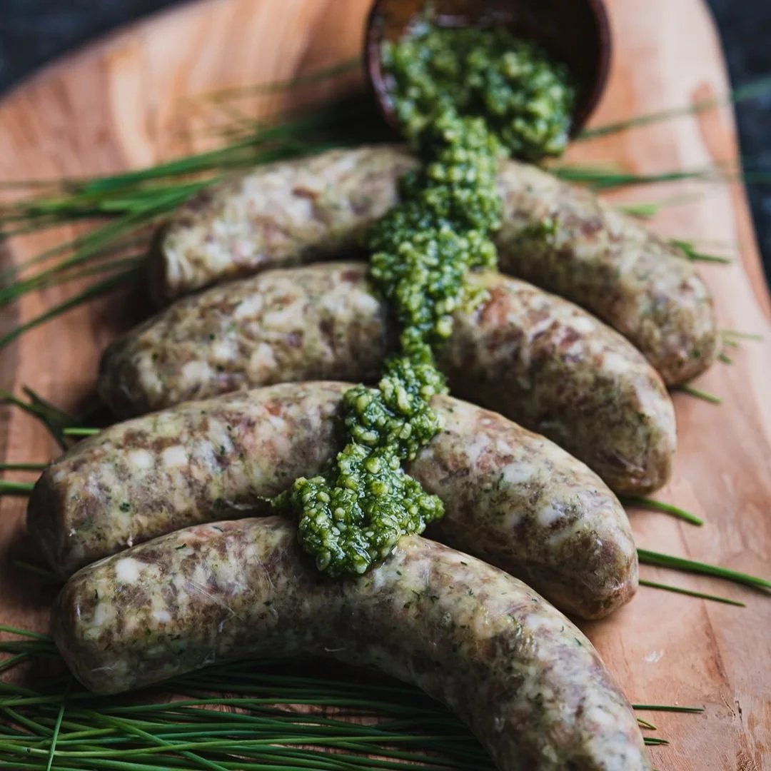Raw sausage links topped with green sauce, garnished with herbs, on a wooden serving board.