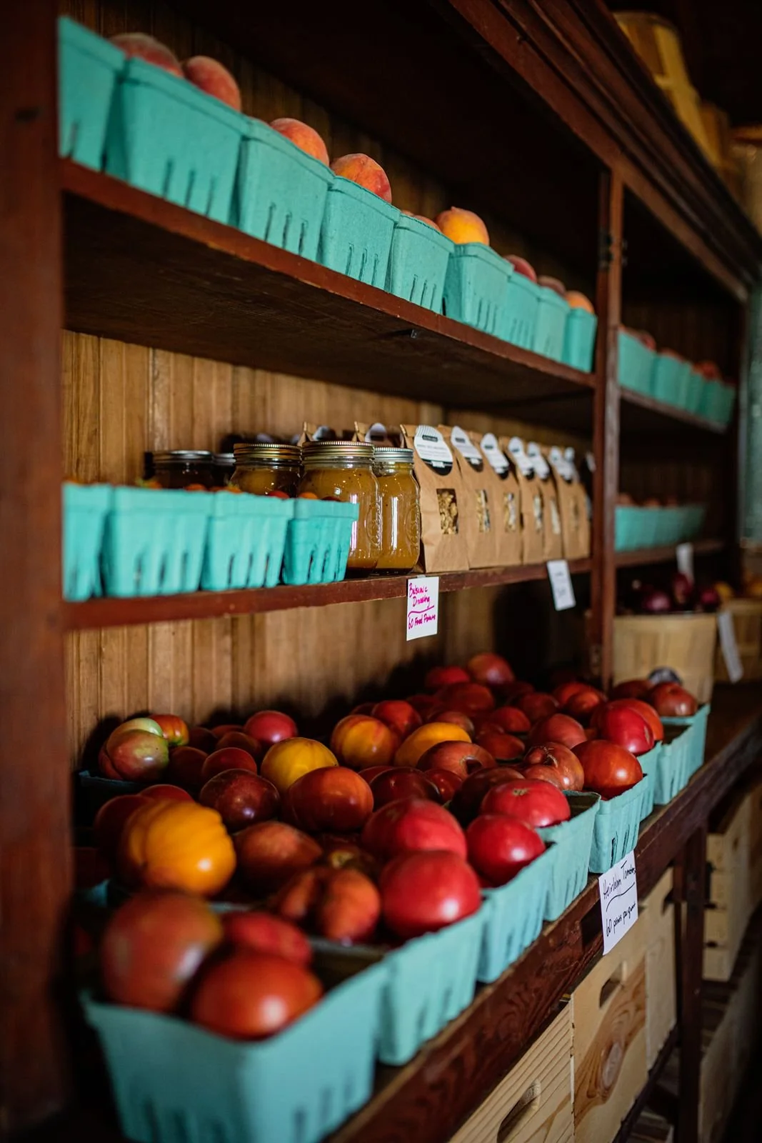 Wooden shelves display baskets of red and green apples, jars of preserved fruits or vegetables, and packaged dried goods, in Moxie Farm's pantry storage.