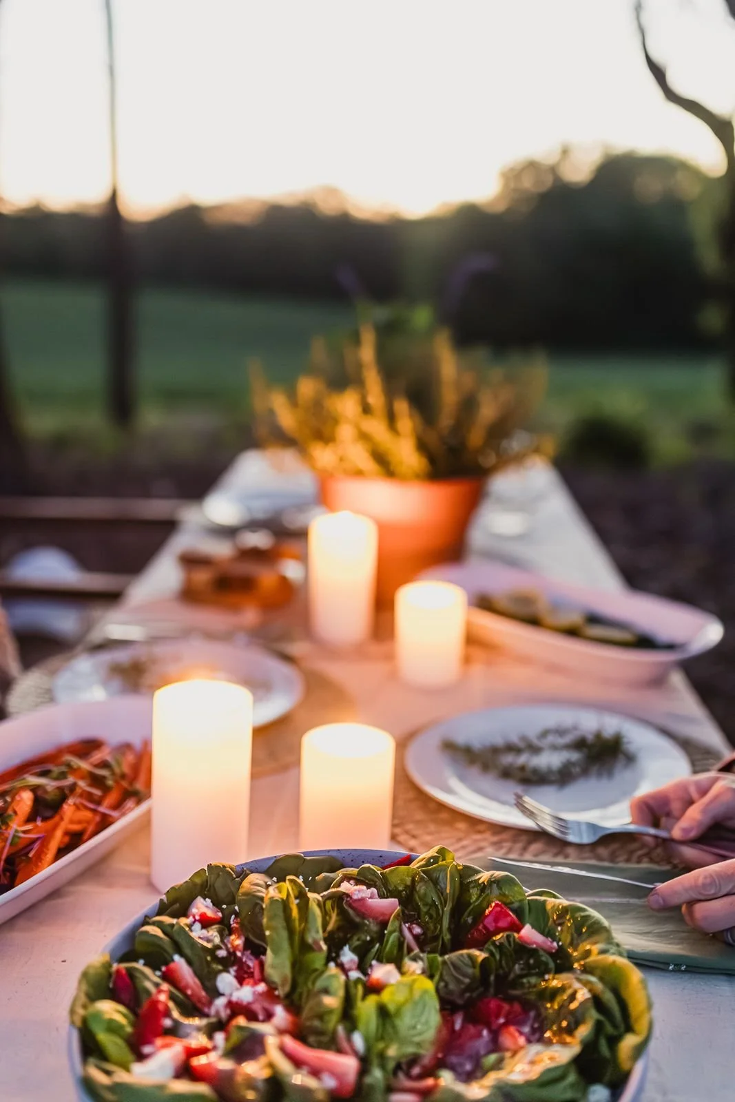 An outdoor dinner table set with salads, candles, plates, and utensils during sunset, with a green landscape in the background.
