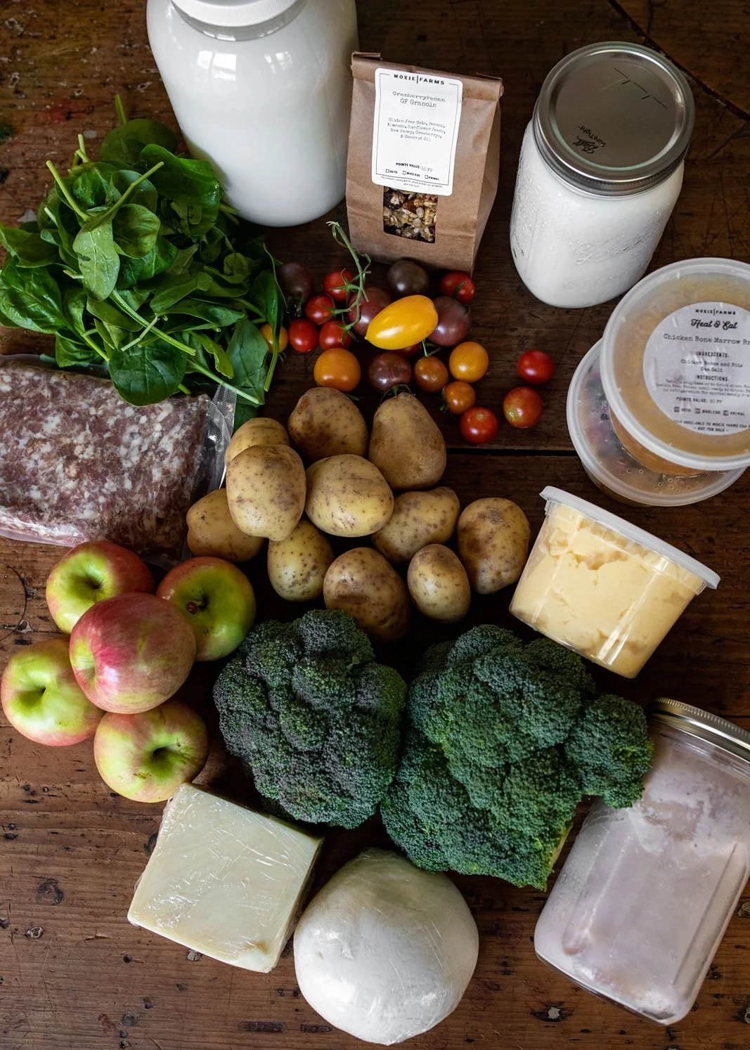 Fresh vegetables, fruits, dairy products, and grains available through Moxie Farm's food membership, including tomatoes, potatoes, broccoli, apples, cheese, milk, and granola.