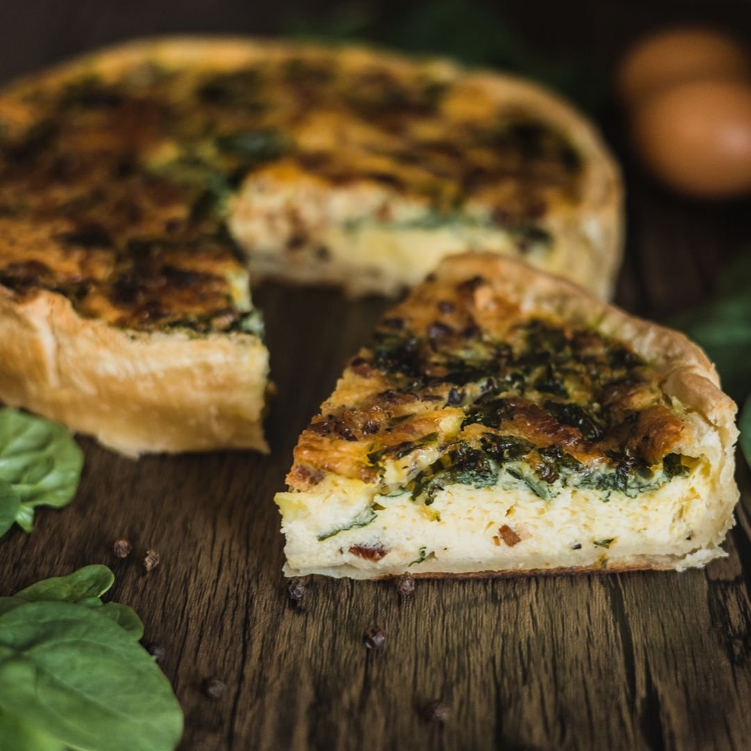 A slice of spinach and cheese quiche on a rustic wooden surface with whole eggs and lettuce leaves around it.