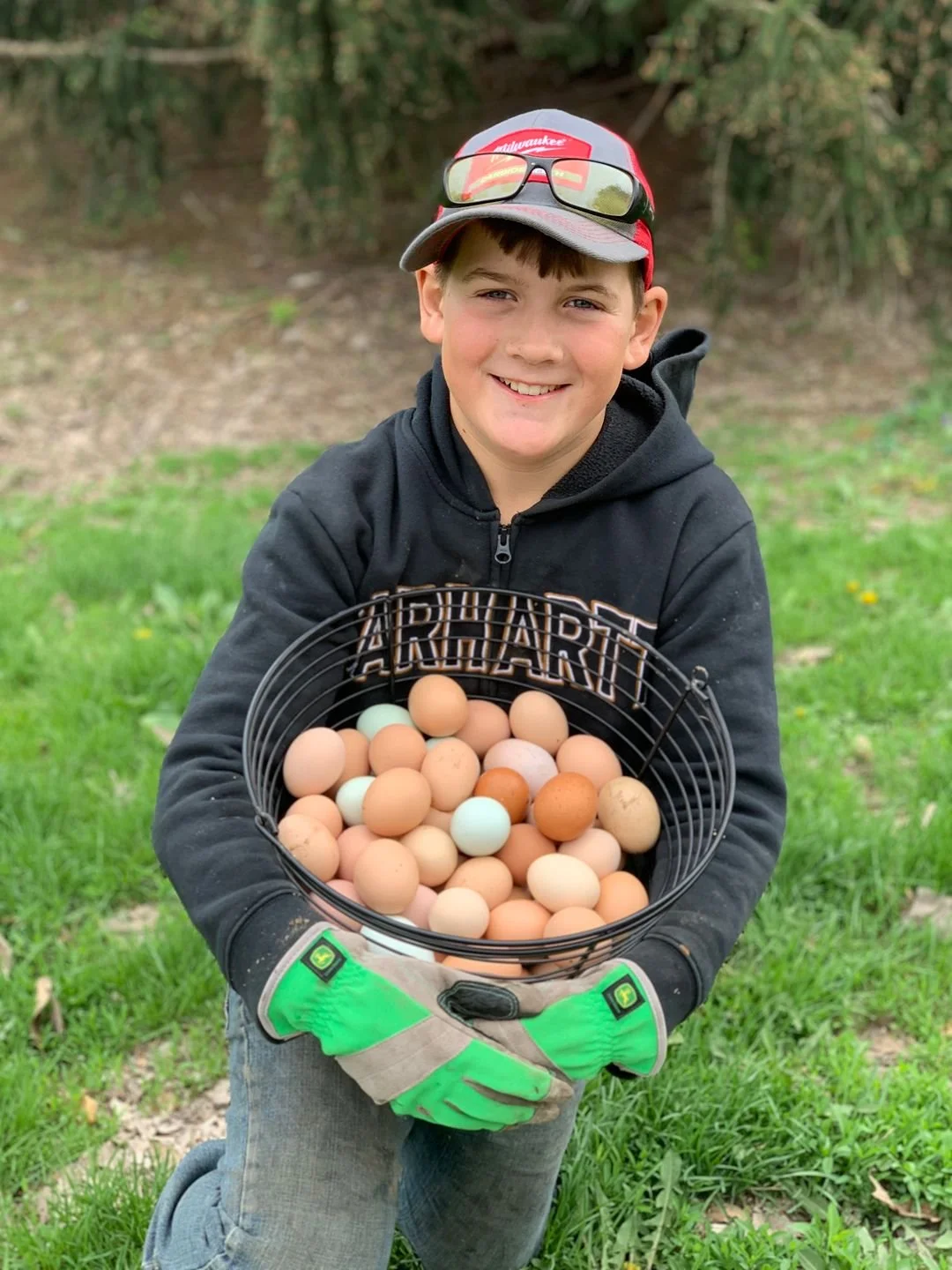 A young boy smiling and holding a basket of eggs outdoors on a grassy area.