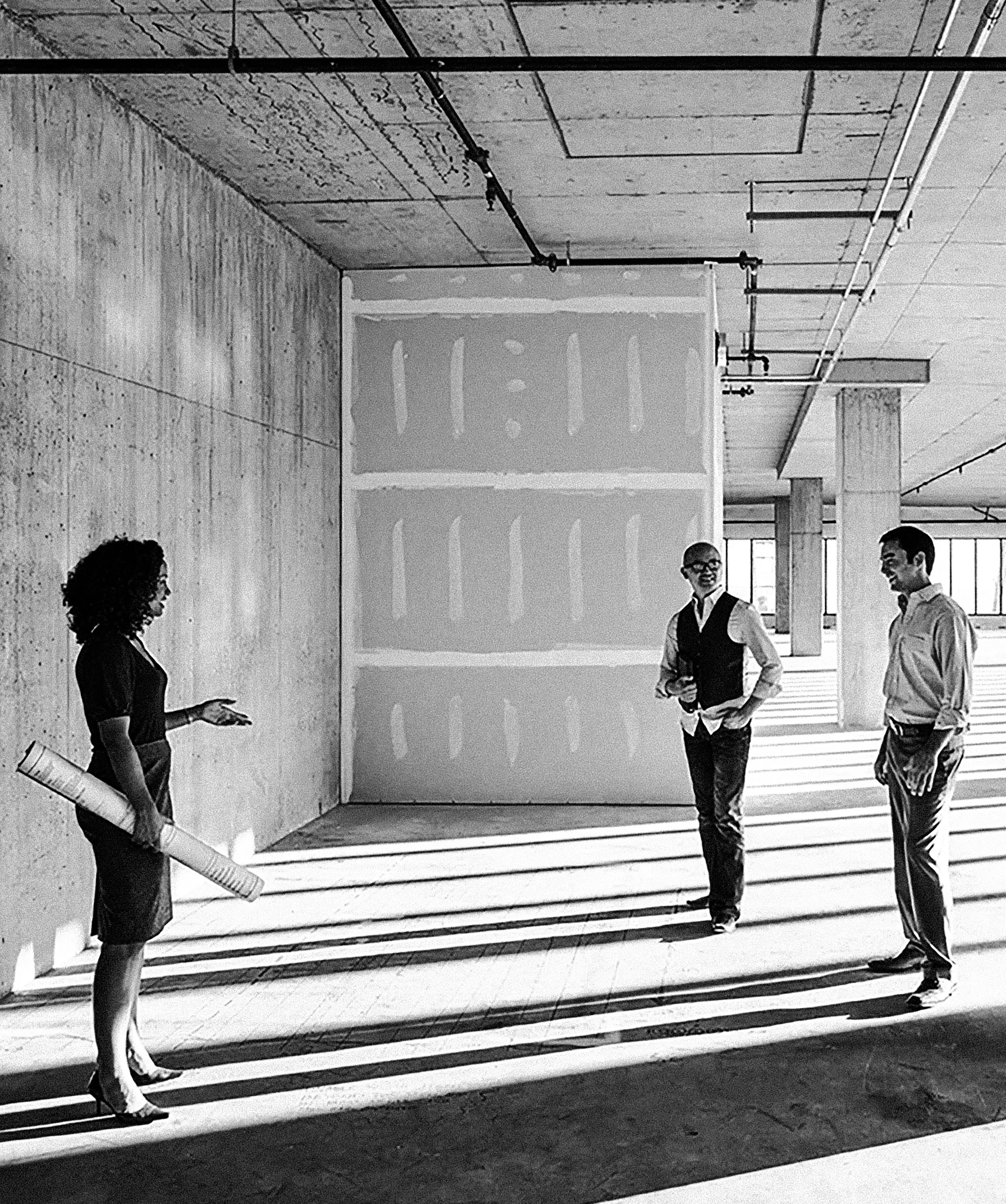 Four people are having a conversation in an industrial or modern office space with exposed concrete walls and large windows. One woman with curly hair and dressed in dark clothing is gesturing with her hand. Two men in business casual attire are standing and smiling, and a woman with glasses and a vest is also smiling, engaged in the conversation.