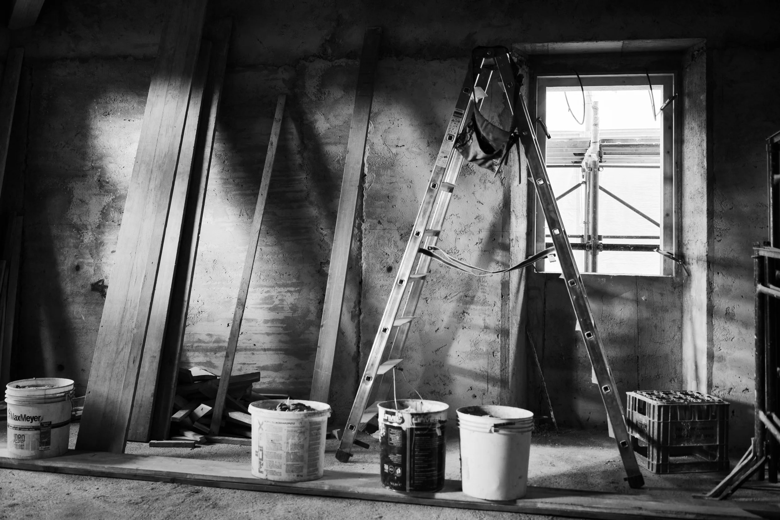 Interior view of a room under construction with wooden framing, an aluminum ladder, buckets of paint or drywall compound, and a window with scaffolding outside.