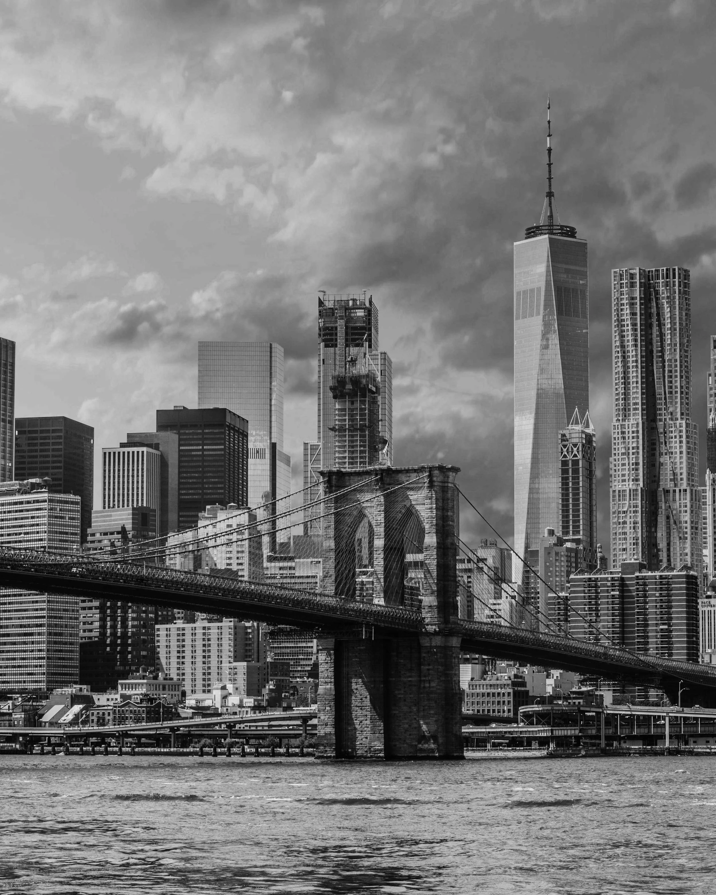 Black and white photo of the New York City skyline with skyscrapers, including One World Trade Center, and a bridge over the river in the foreground.