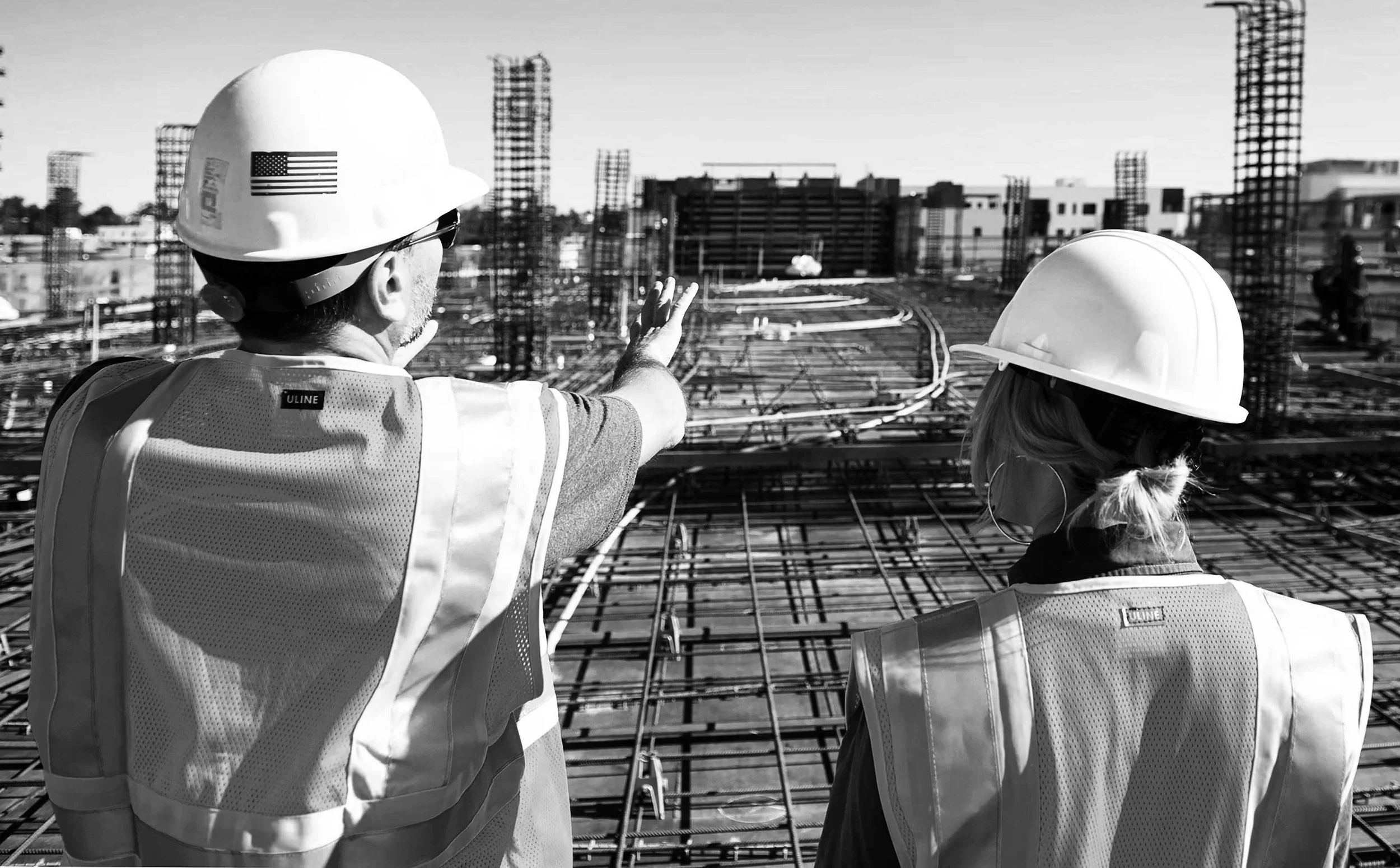 Two construction workers wearing hard hats and safety vests observing a building site with steel framework.