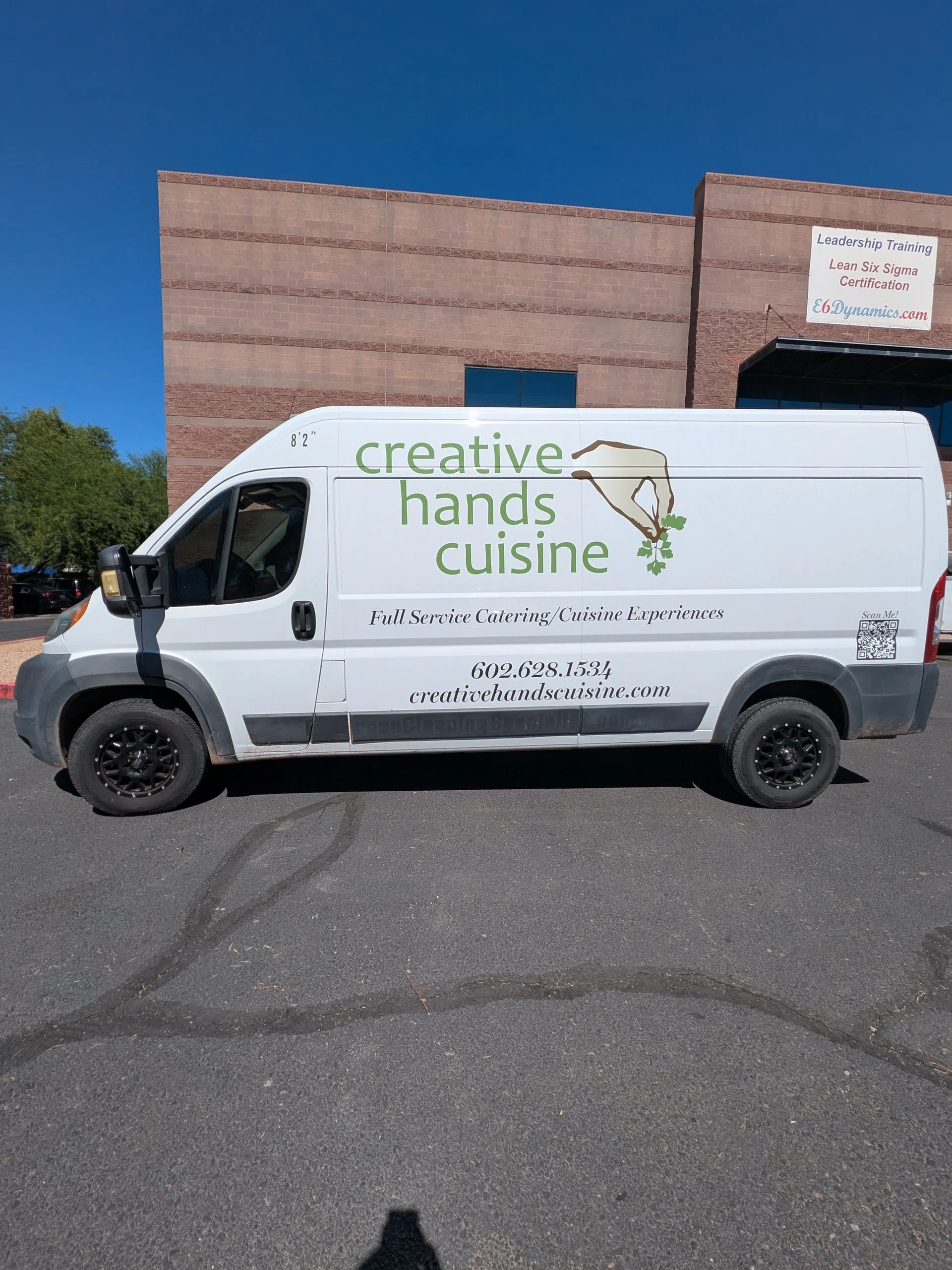 A white van parked in front of a red brick building with a sign advertising leadership training and Lean Six Sigma certification from E&N Dynamics. The van has a logo of a hand holding a sprouting plant with the text 'creative hands cuisine' and cont
