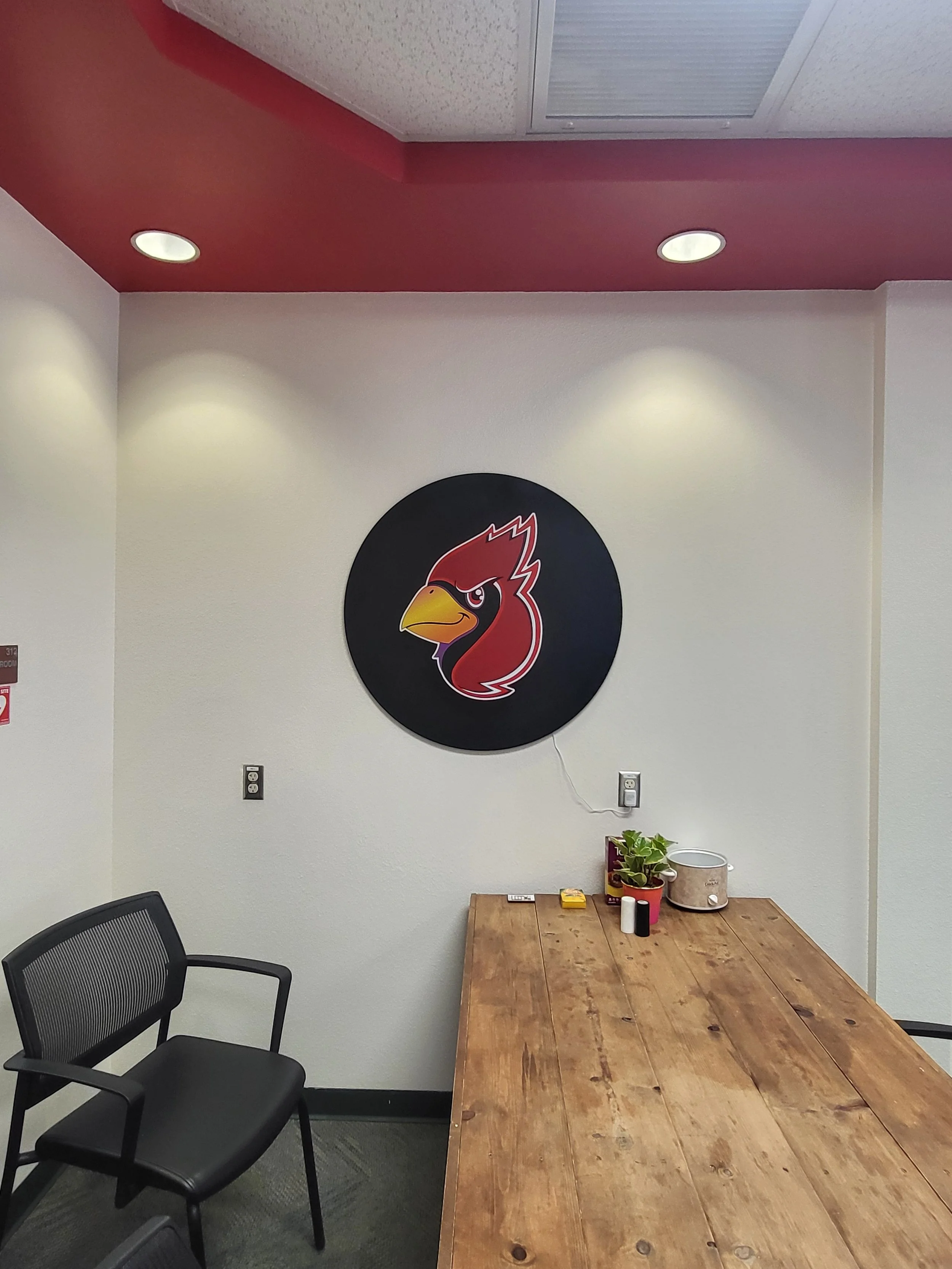An office conference room with a wooden table, black office chair, a wall with a logo of a red bird, plants and items on the table, ceiling lights, and an electrical outlet.