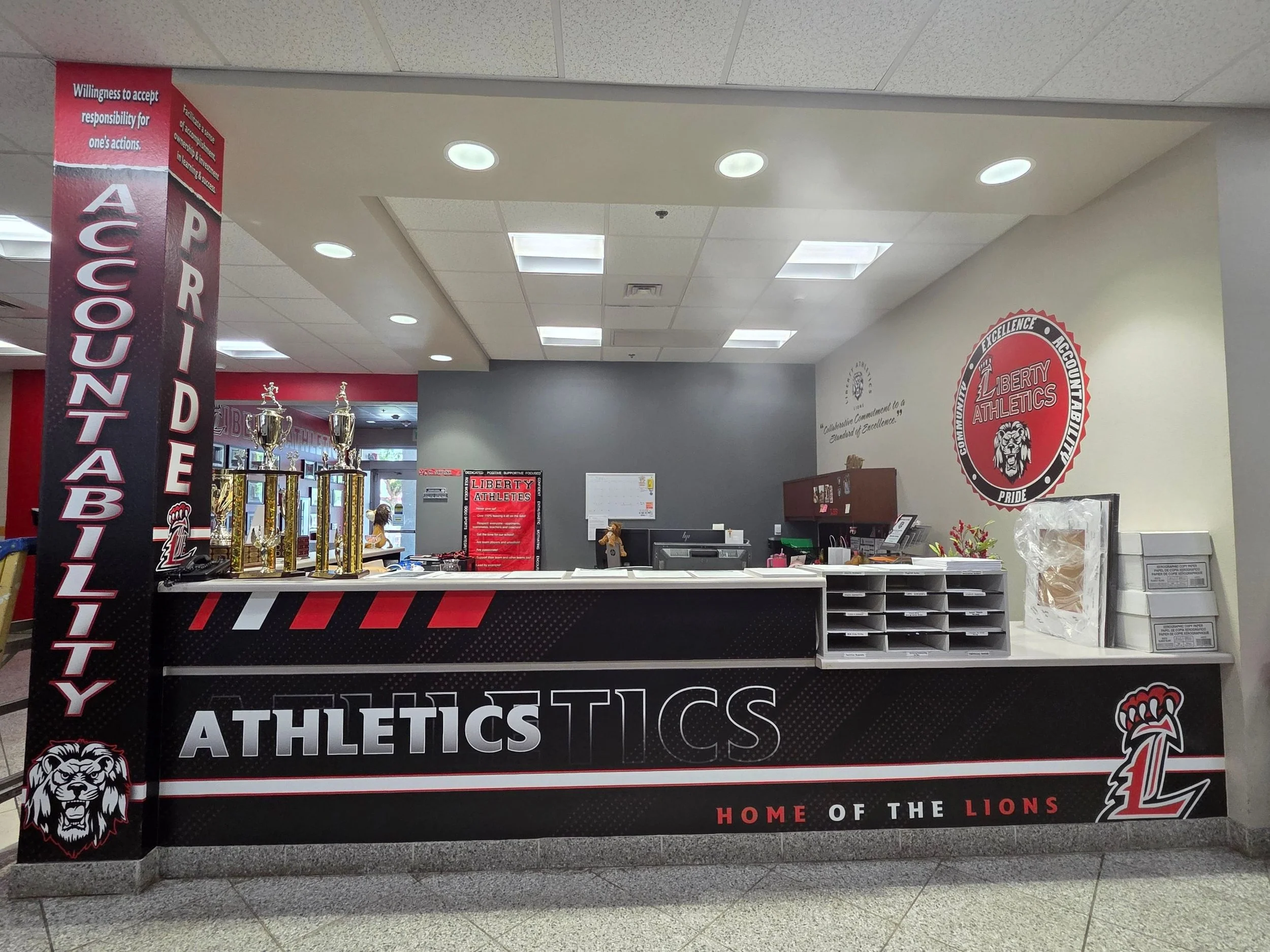 School reception desk with trophies, posters, and signage for Liberty Athletics and the Great Lions mascot.
