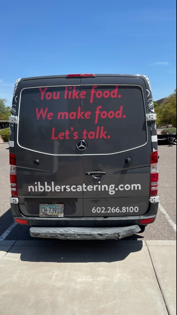 Black catering van with signs in red and white text. Red text says: 'You like food. We make food. Let's talk.' White text displays website 'nibblerscatering.com' and phone number '602.266.8100'. The van has a Arizona license plate and is parked in a 