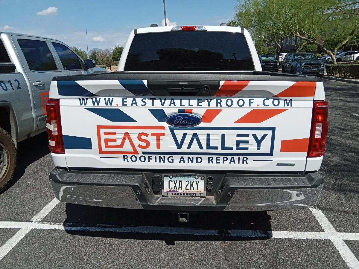 The back of a Ford pickup truck with a business advertisement for East Valley Roofing and Repair, including the website URL www.eastvalleyroof.com, the license plate CXA 2KY, parked in a parking lot with other vehicles and trees in the background.