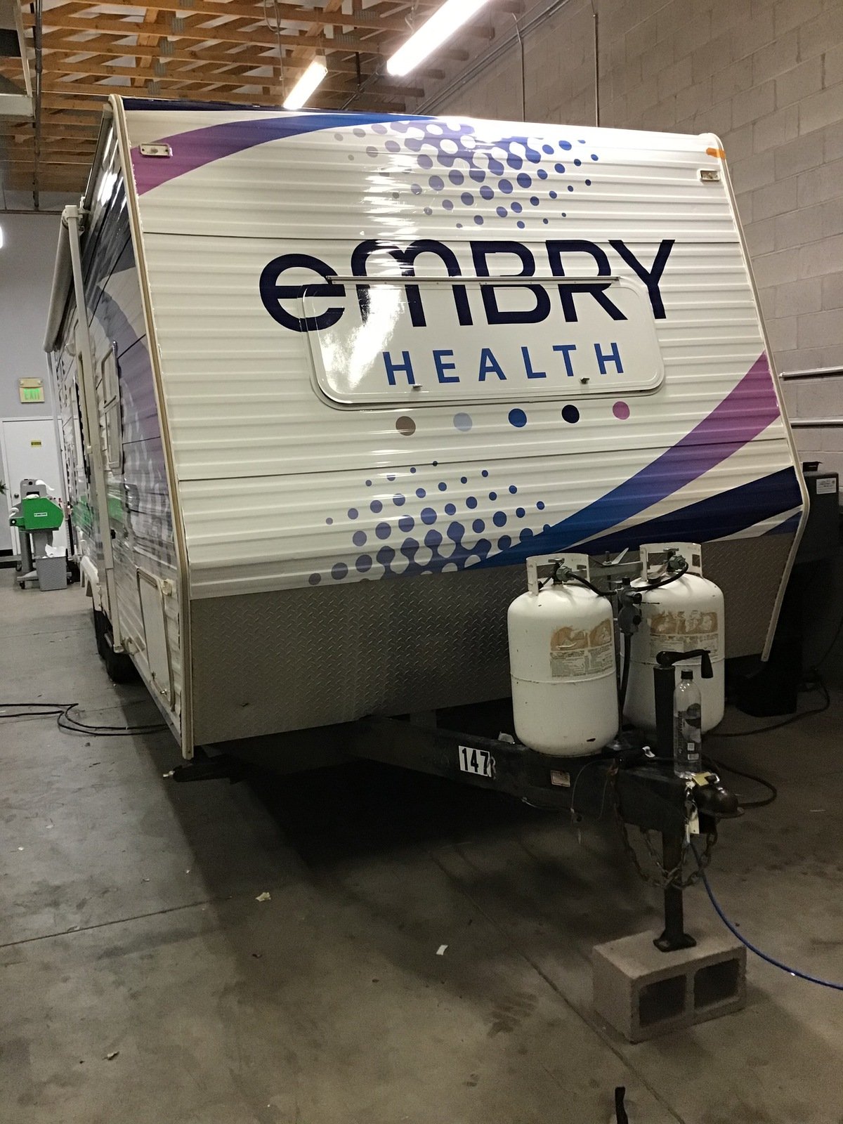 A trailer with 'Embury Health' written on the side, parked indoors, with propane tanks and cleaning supplies in front.