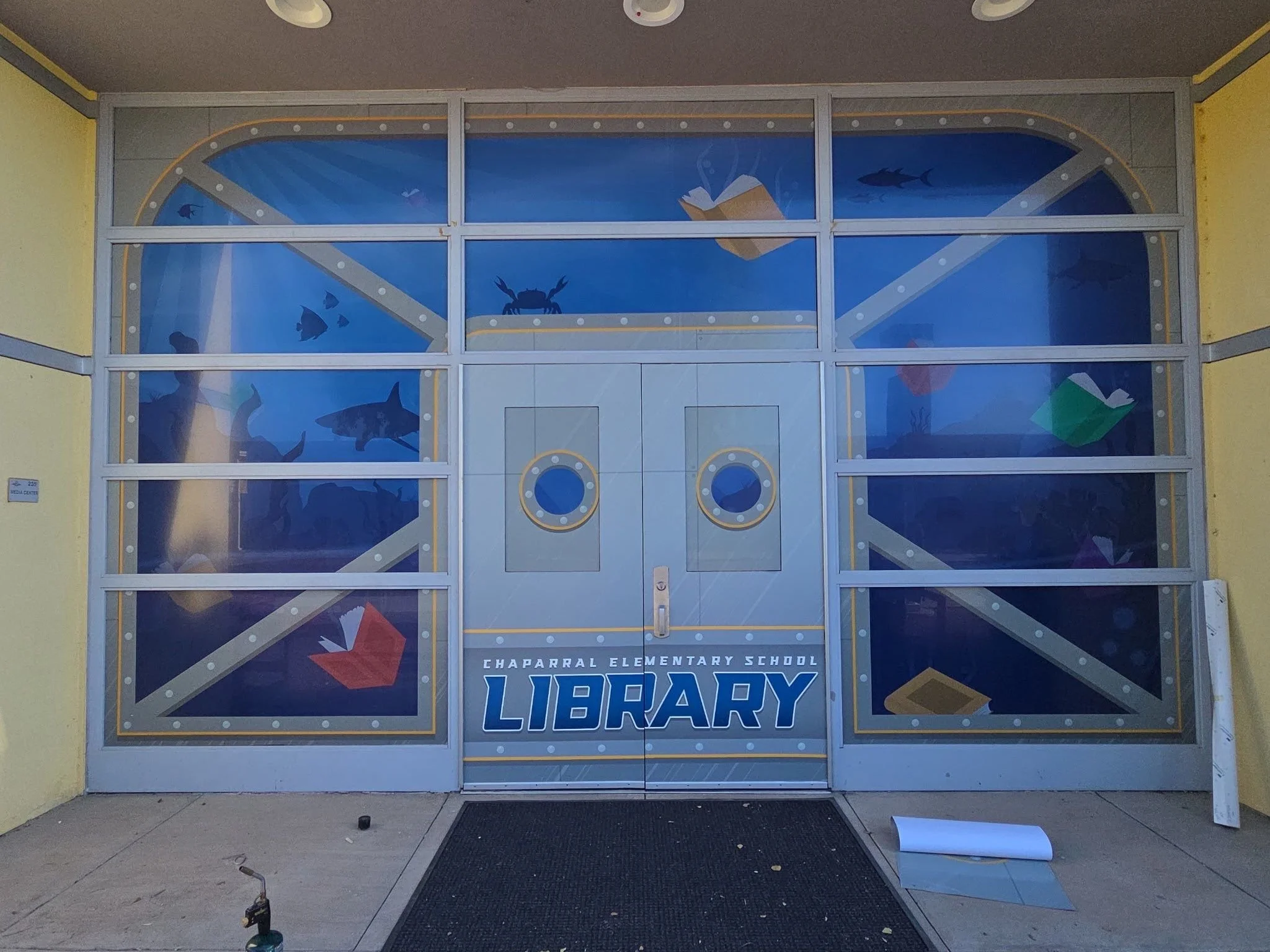 Front view of a school entrance with a colorful mural featuring underwater sea creatures and books, and the text 'Chaparral Elementary School Library' on the door.