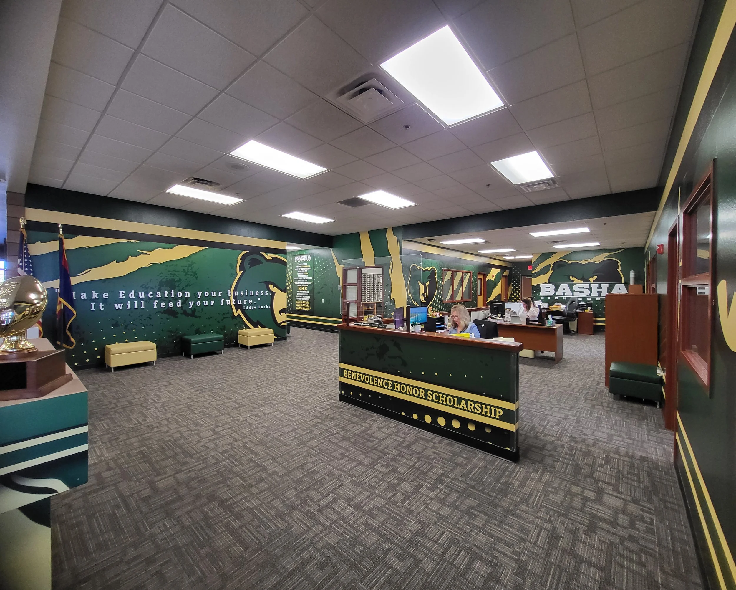 School reception area with a mural of a bear and green trees on the wall, a desk with computers, and people working.